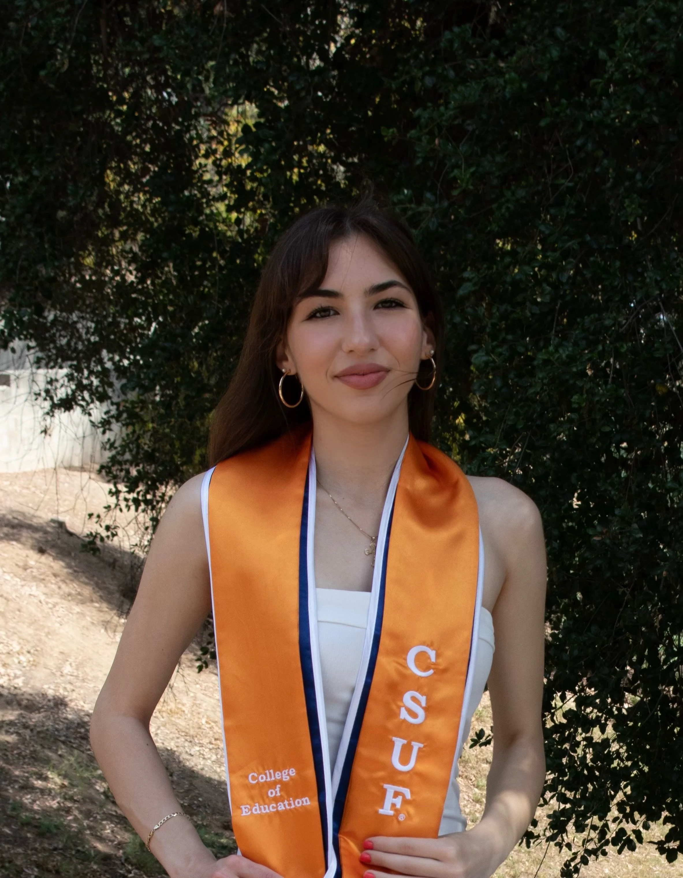 Young woman at graduation ceremony wearing an orange stole with 'CSUF' and 'College of Education' embroidered on it, standing outdoors with trees in the background.