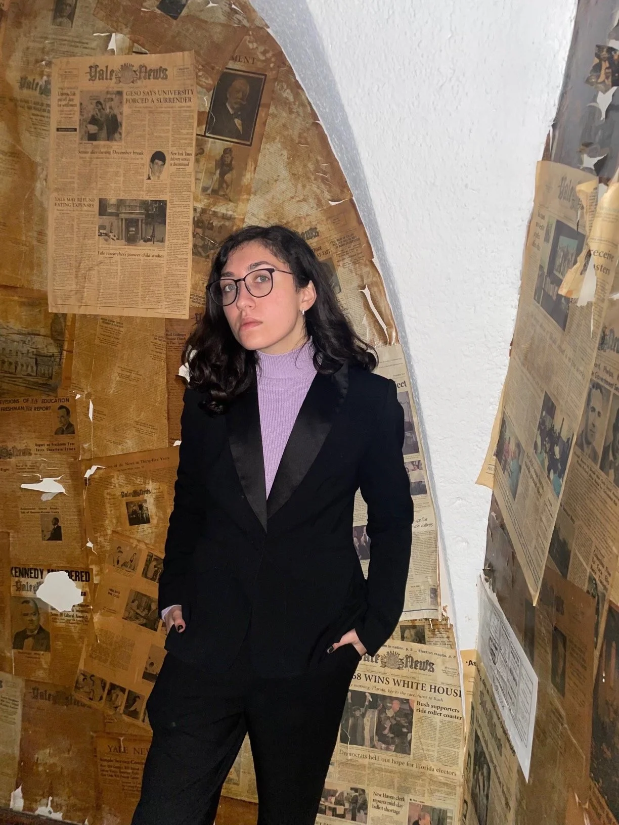 Young woman with glasses and dark curly hair wearing a black blazer over a purple turtleneck, standing in a corner with walls covered in peeling yellowed newspapers.