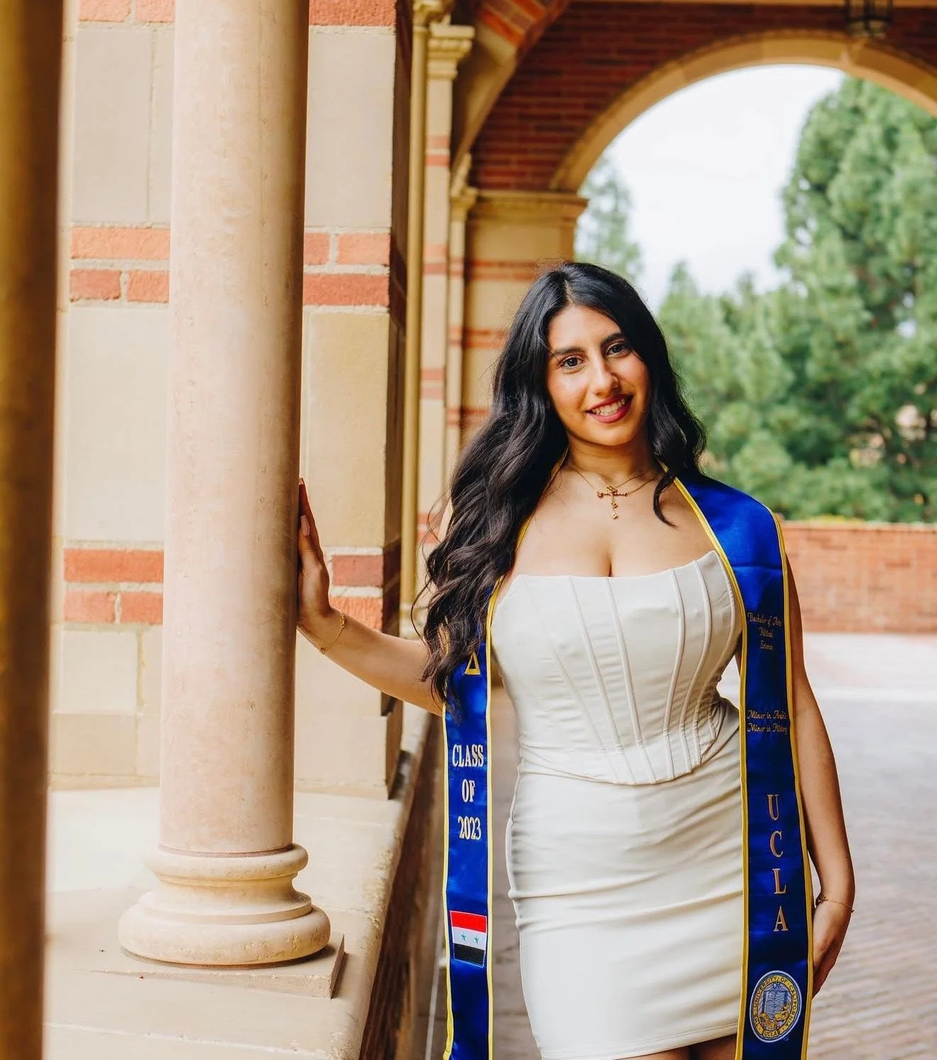 A young woman in a white dress and a blue graduation sash with gold trim, standing outdoors by a stone column, smiling at the camera.
