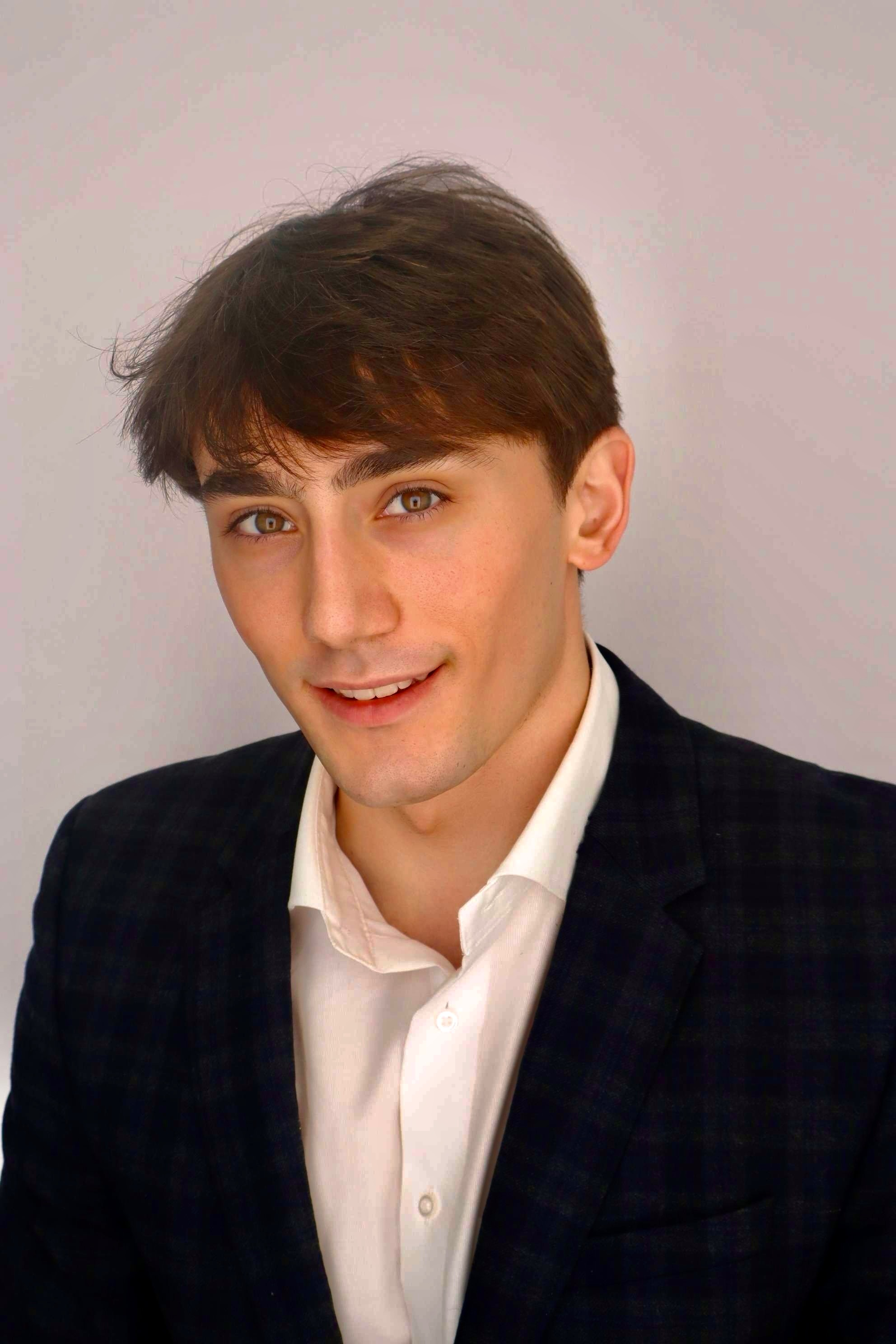 Portrait of a young man with brown hair, wearing a white shirt and dark blazer, smiling at the camera against a plain background.