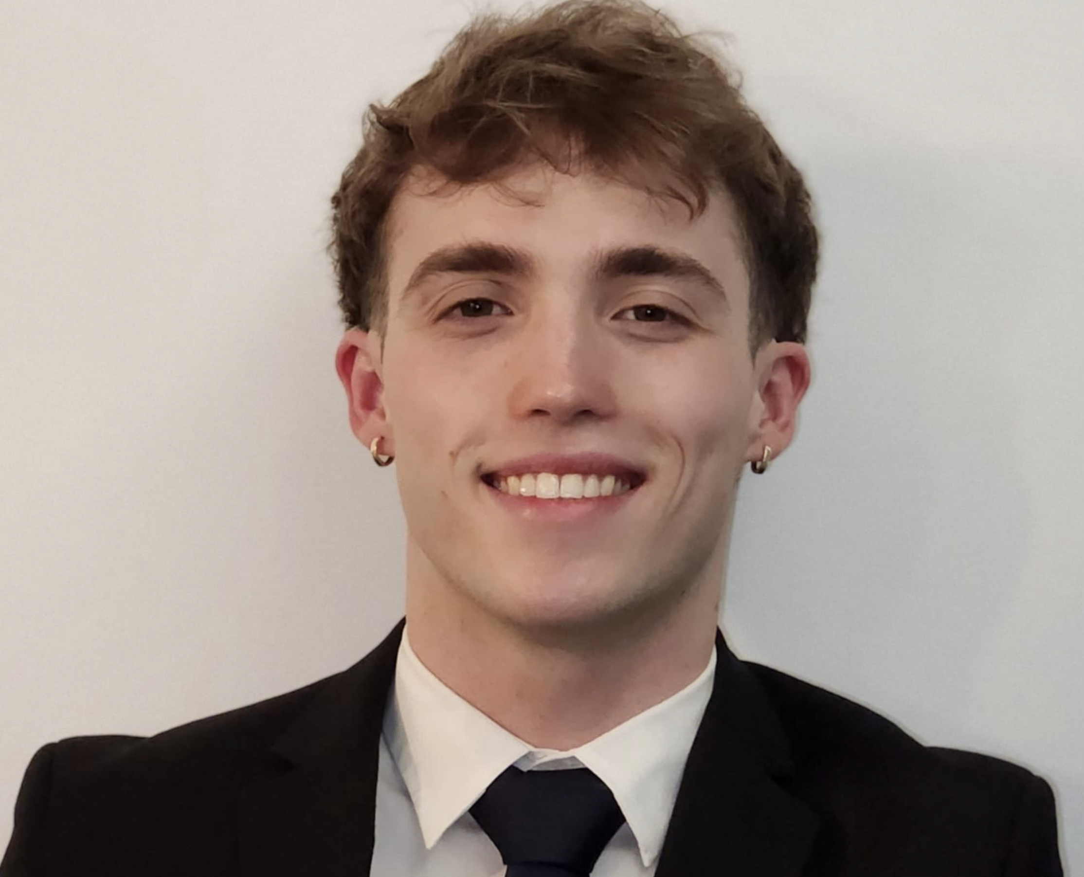 Young man with short, wavy brown hair, wearing a dark suit, white shirt, and black tie, smiling with visible teeth, against a plain white background.