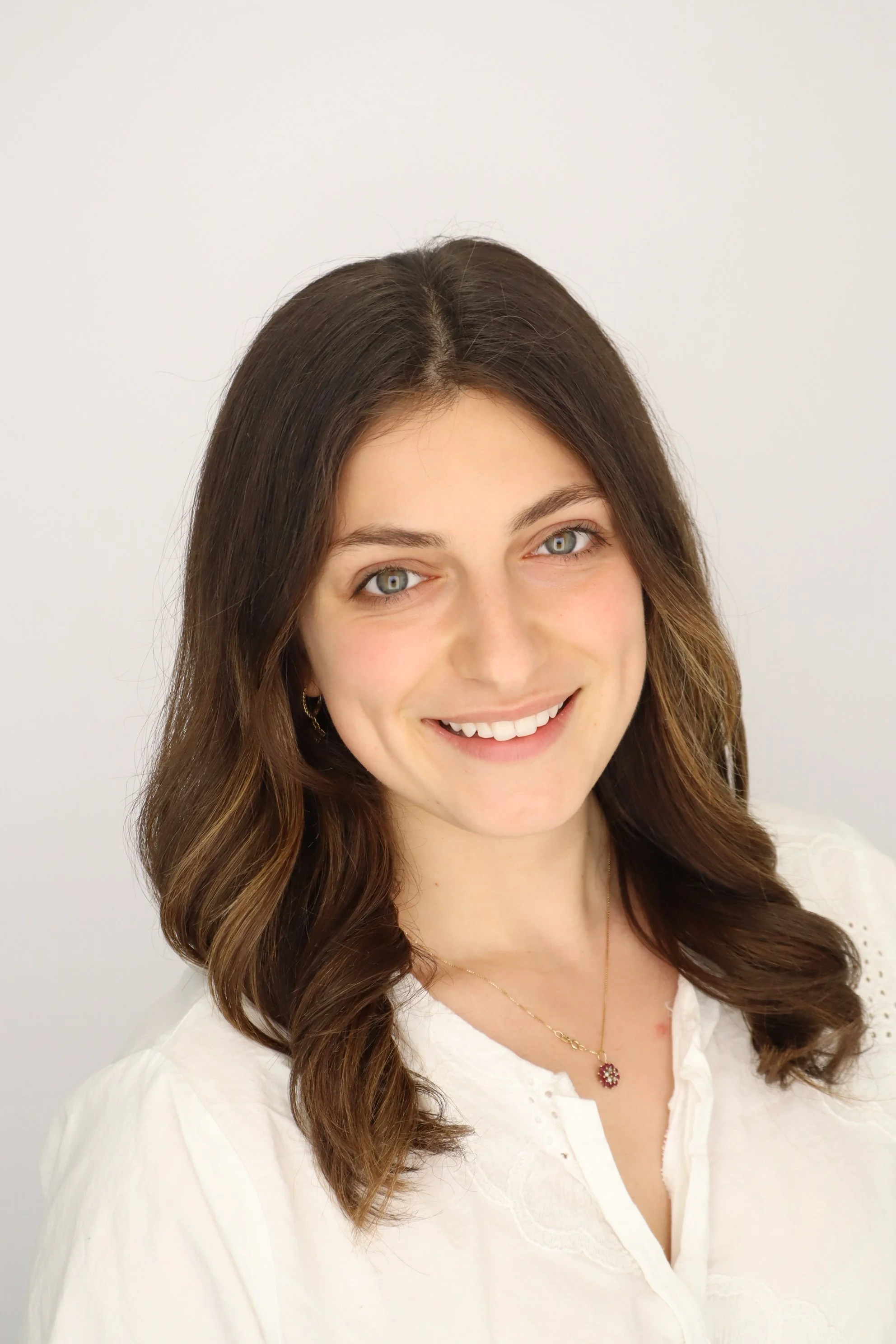 Portrait of an Ivy League tutor, young woman smiling, with wavy brown hair, blue eyes, wearing a white top and a delicate necklace with a pendant, against a plain white background.