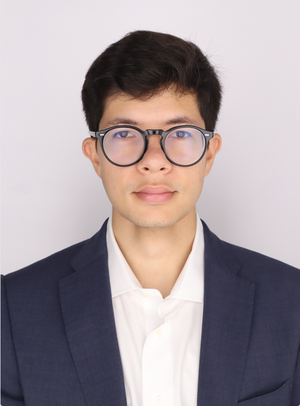 A young man with dark hair wearing round glasses, a white button-up shirt, and a navy blazer, posing against a plain light background.