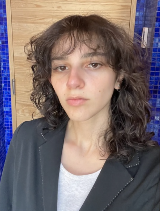 A young person with curly brown hair, wearing a black blazer over a white shirt, taking a selfie indoors with wooden and blue tiled walls in the background.