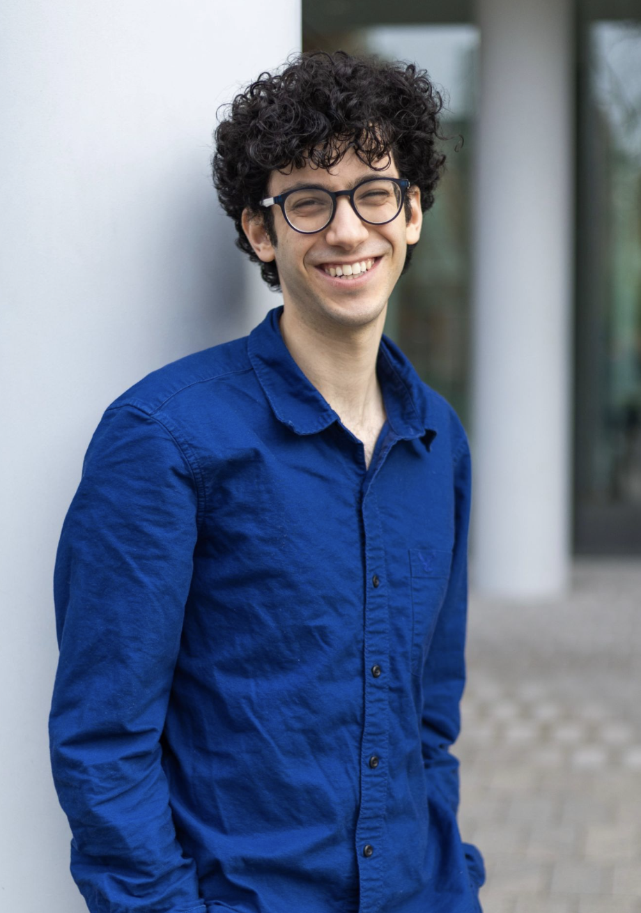A young man with curly dark hair and glasses smiling while leaning against a white wall outdoors.