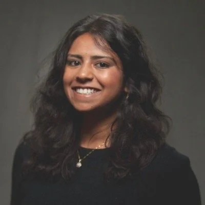 A young woman with dark, wavy hair smiling, wearing a black top and a necklace, against a dark background.