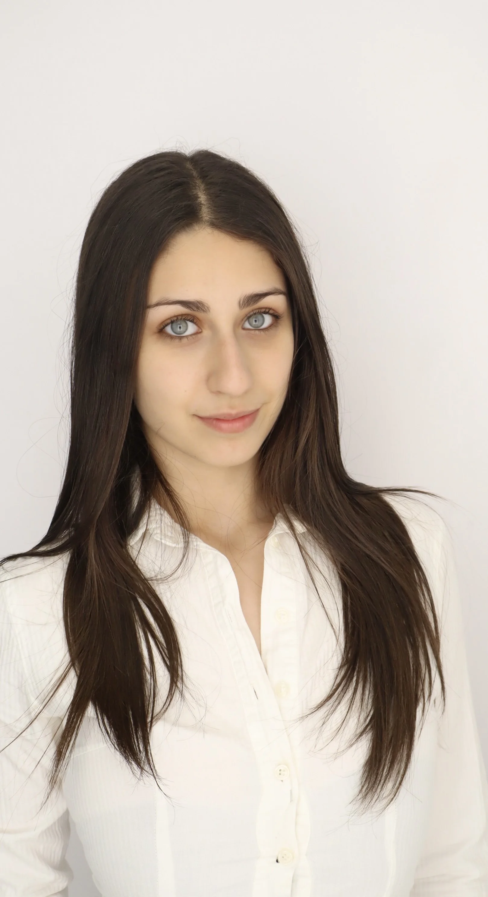 A young woman with long brown hair and blue eyes, wearing a white button-up shirt, standing against a plain white background.
