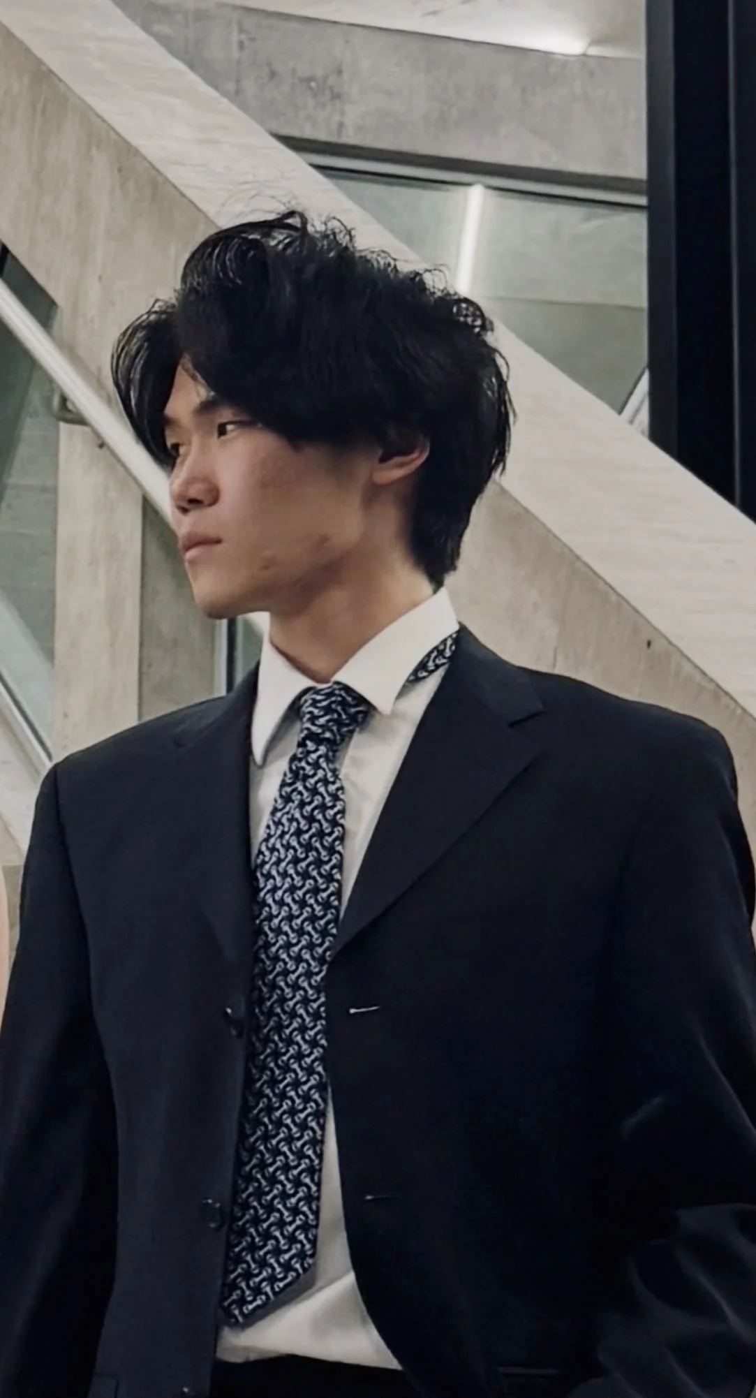 A young man with black wavy hair wearing a black suit and a patterned tie, standing indoors in front of a concrete wall and stairs.