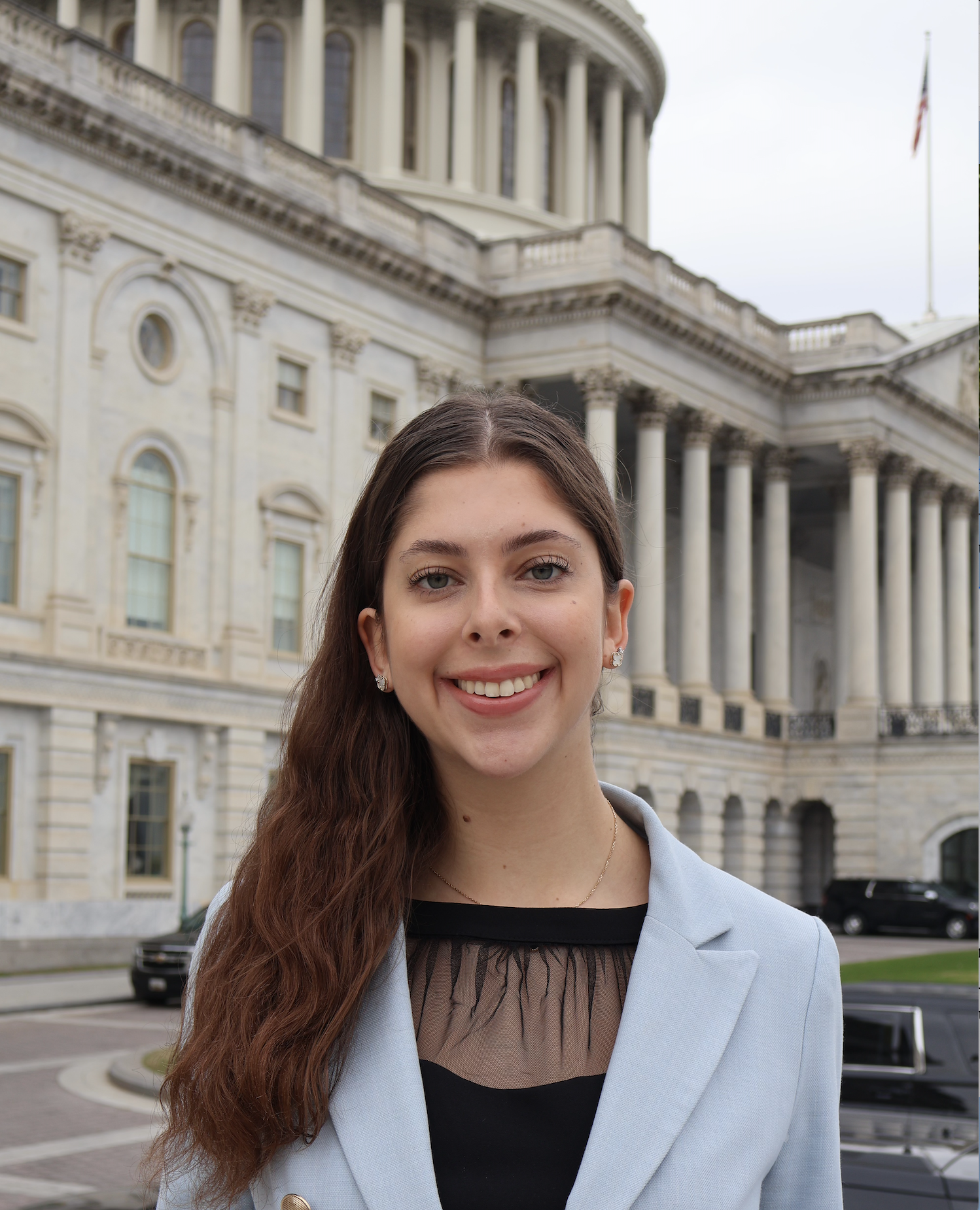 A young woman with long brown hair and earrings, smiling, standing outside near a government building with classical architecture, columns, and an American flag.