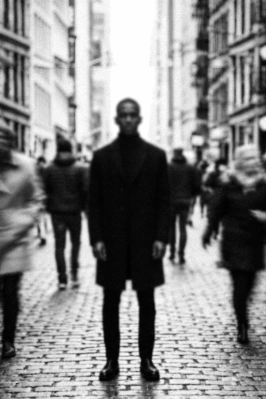 Black and white photo of a man wearing dark clothing and glasses standing in the middle of a busy city street lined with tall buildings and people walking.