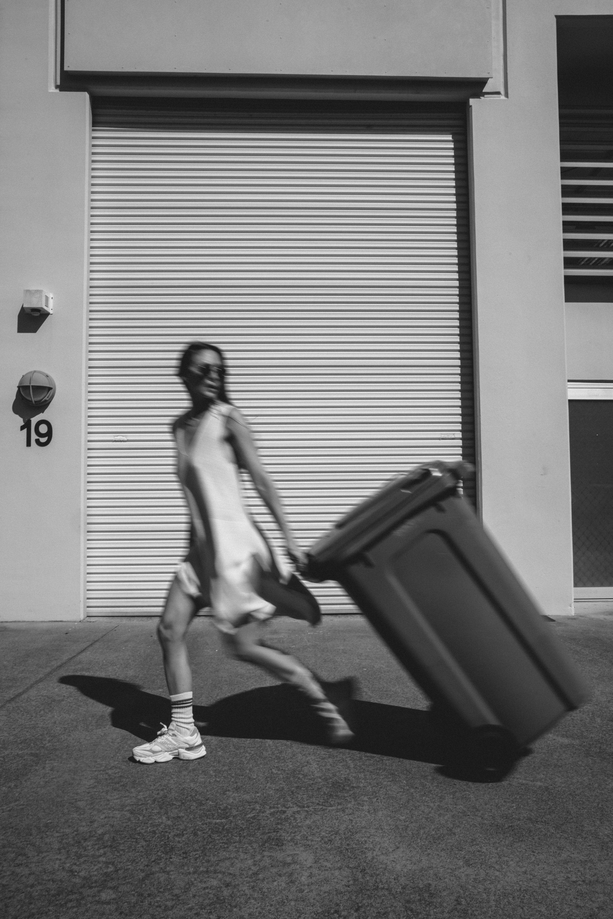 A woman with long hair, wearing sunglasses, a light-colored dress, and sneakers, is pulling a rolling suitcase on a sidewalk in front of a building with a large metal garage door.