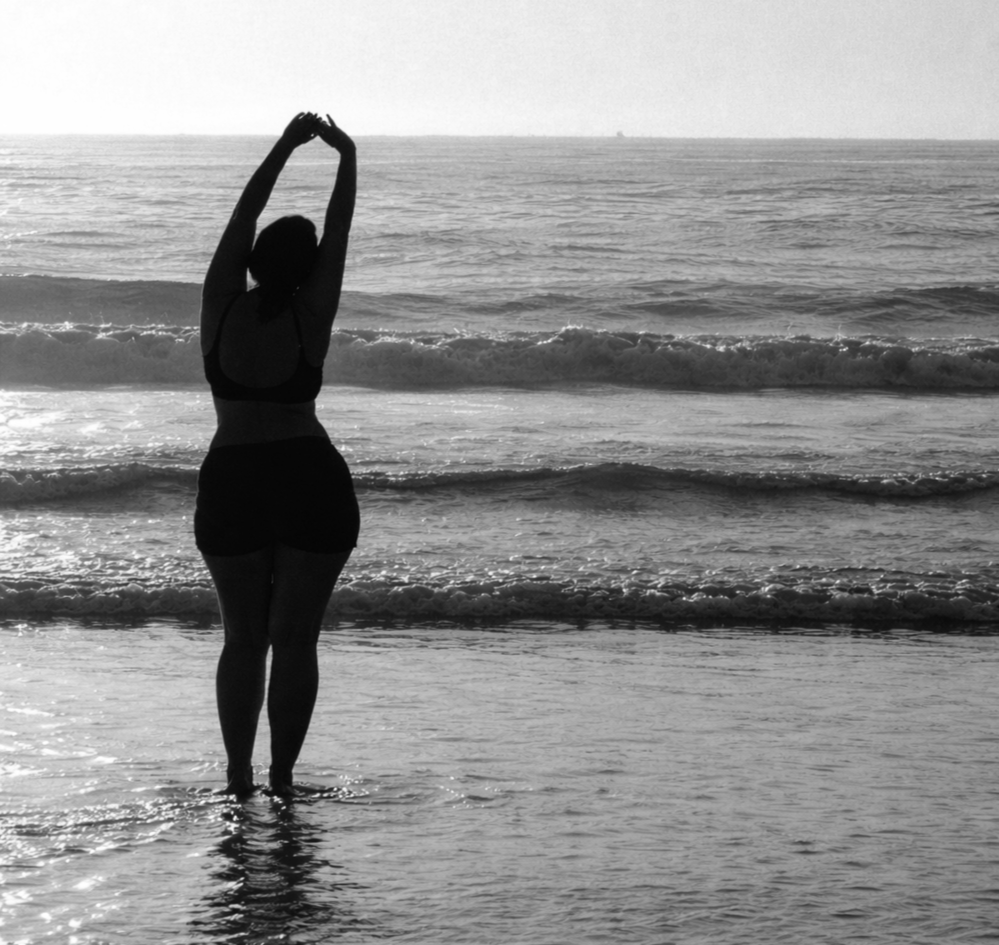 A person in a swimsuit standing in shallow water at the beach, stretching with arms raised, facing the ocean at sunset or sunrise.