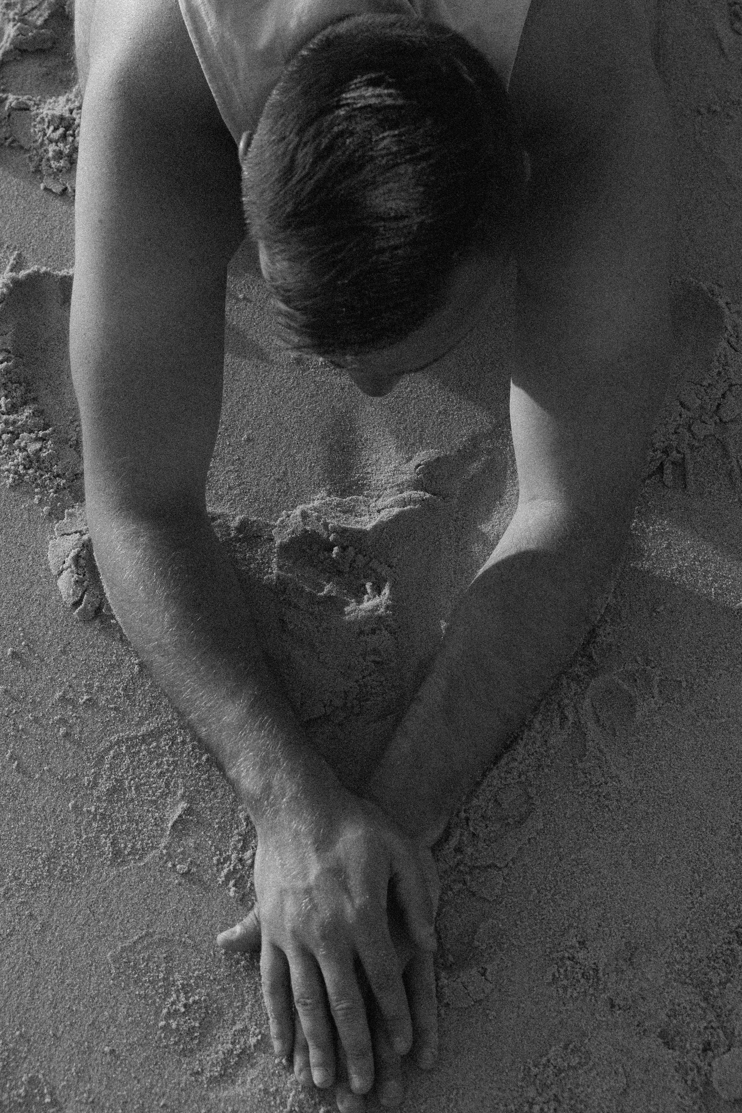 A man with slicked-back hair, shirtless, digging a hole in the sand at the beach.