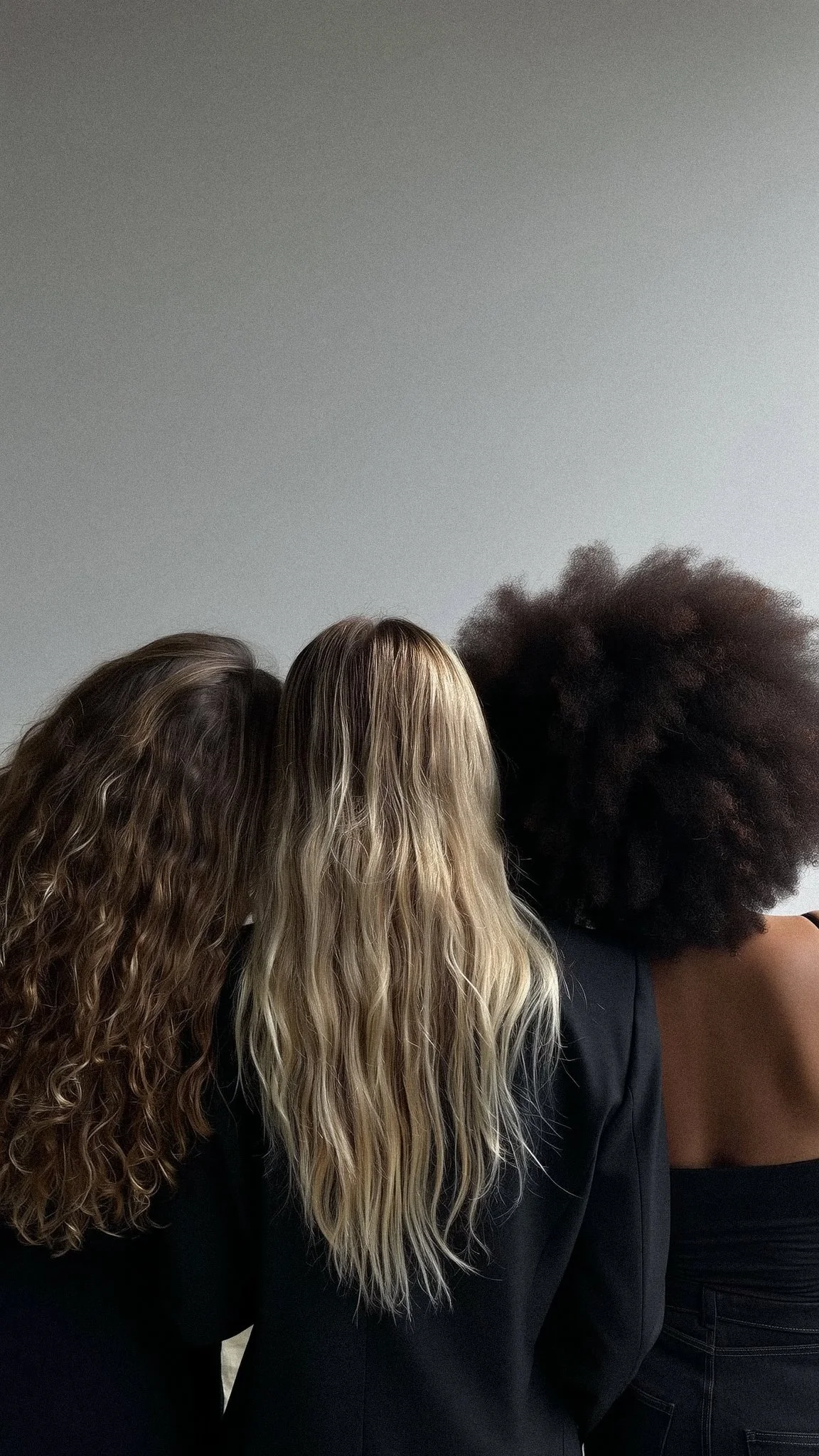Three women with different hair textures standing side by side with backs facing the camera, against a plain gray wall.