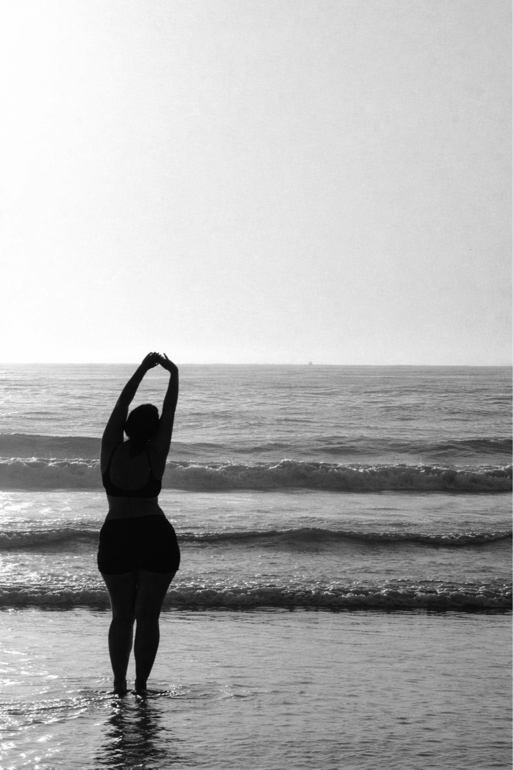 A woman standing in the shallow waters of the ocean at the beach, stretching with her arms raised above her head, silhouetted against the horizon.