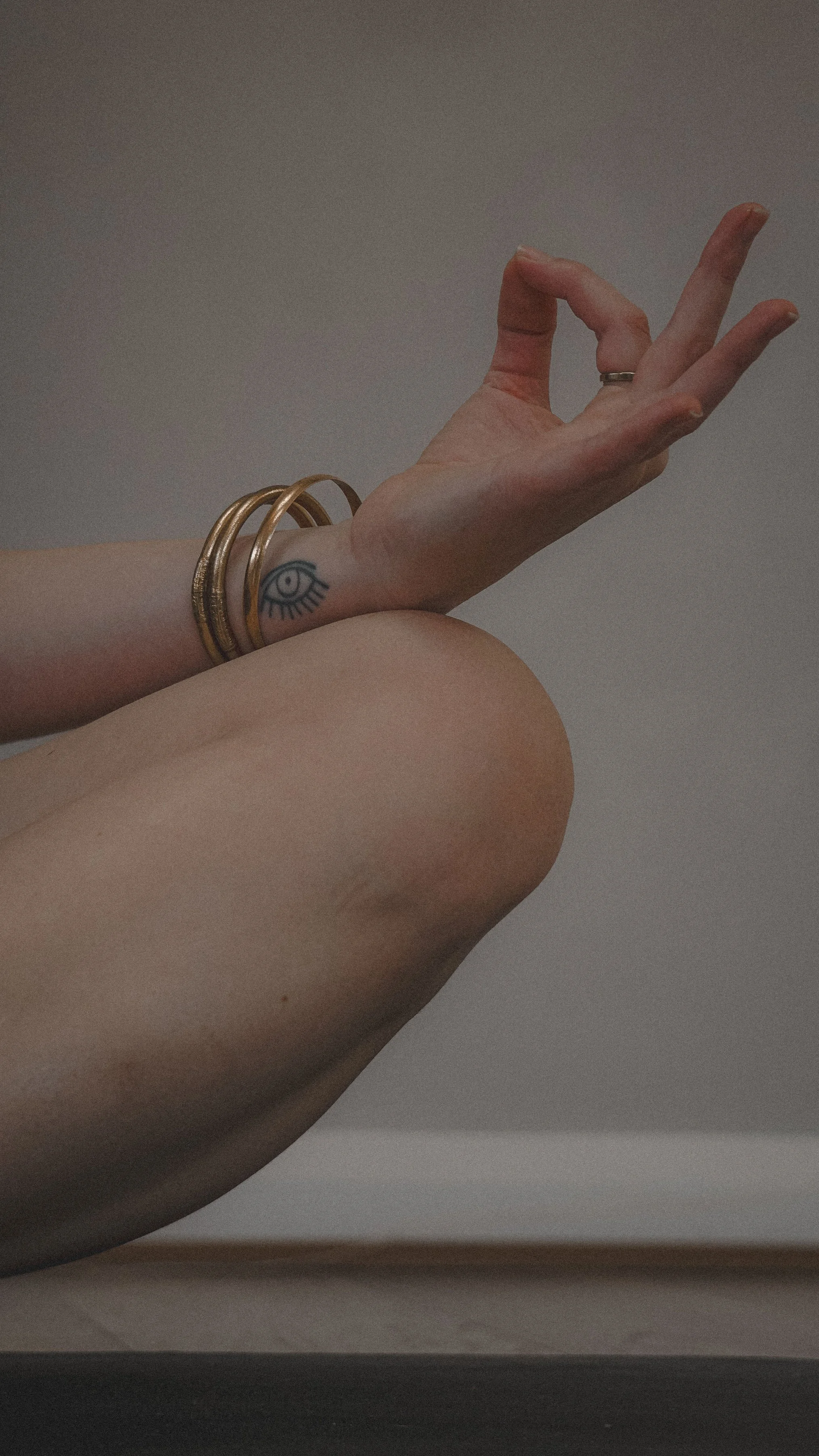 Close-up of a person's arm and knee with minimal skin visible, showing a tattoo of an eye on the wrist and gold bangles, with the hand making an OK gesture.