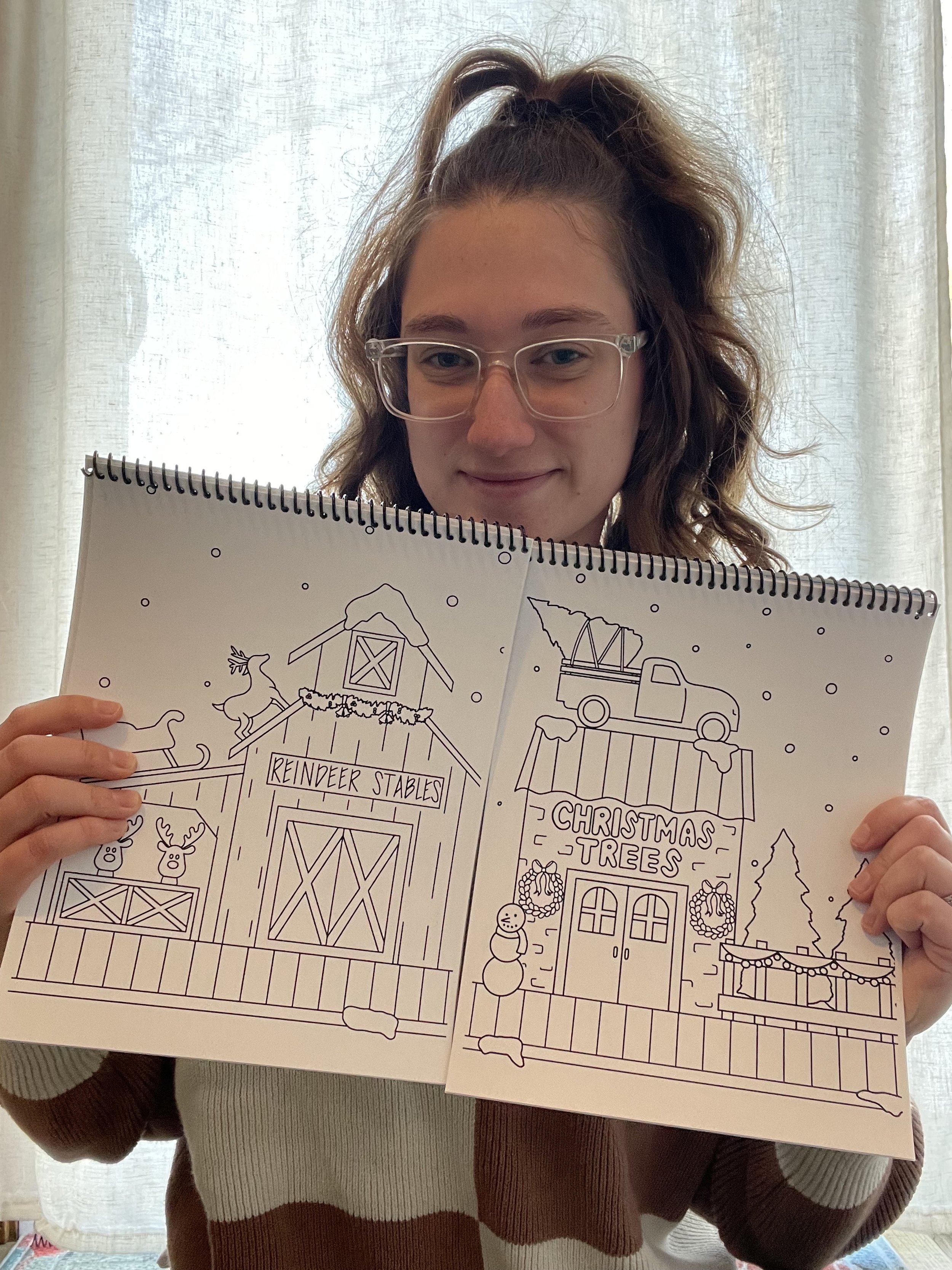 A person with glasses and wavy hair in a ponytail holds up a coloring book showing a Christmas-themed scene, including a reindeer stable and a Christmas tree shop, with snow falling in the background.