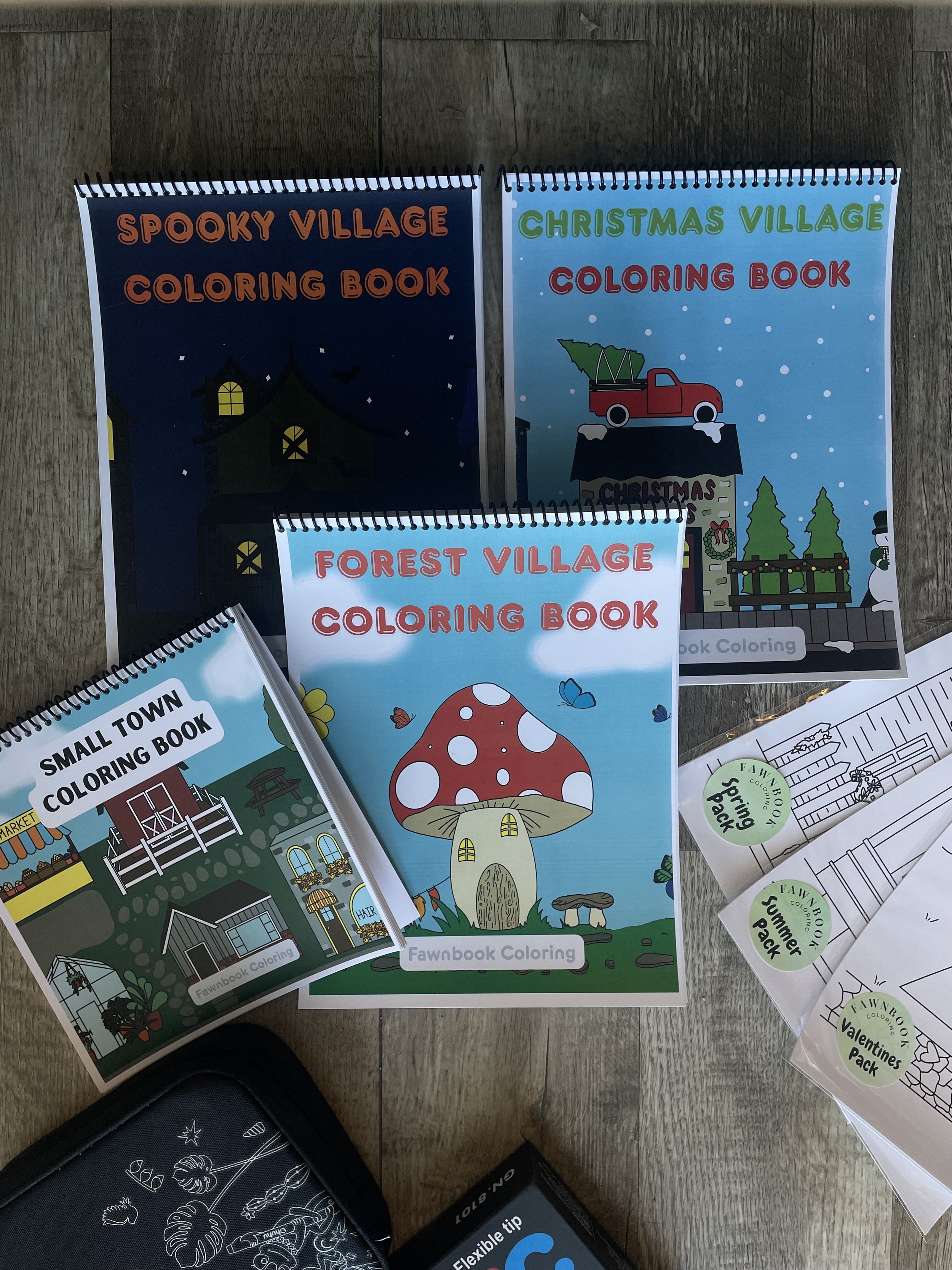 Four children's coloring books with colorful covers on a wooden surface, including 'Spooky Village', 'Christmas Village', 'Forest Village', and 'Small Town'.