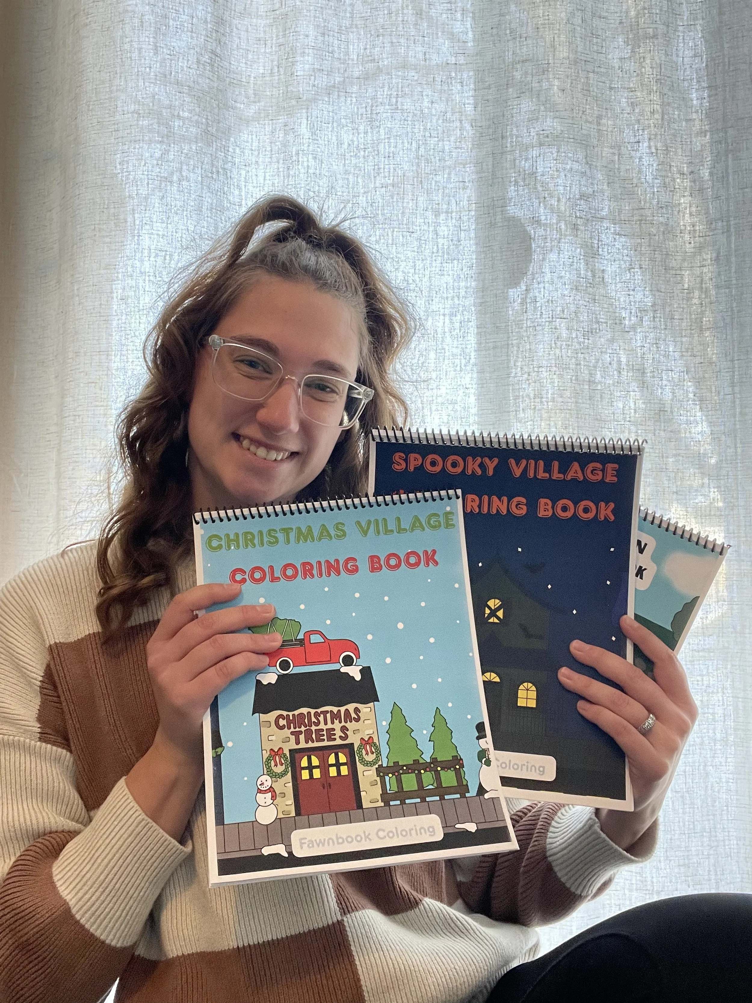 A young woman with glasses smiling and holding three coloring books with holiday-themed covers, including Christmas village, spooky village, and winter village, in front of a sheer curtain.