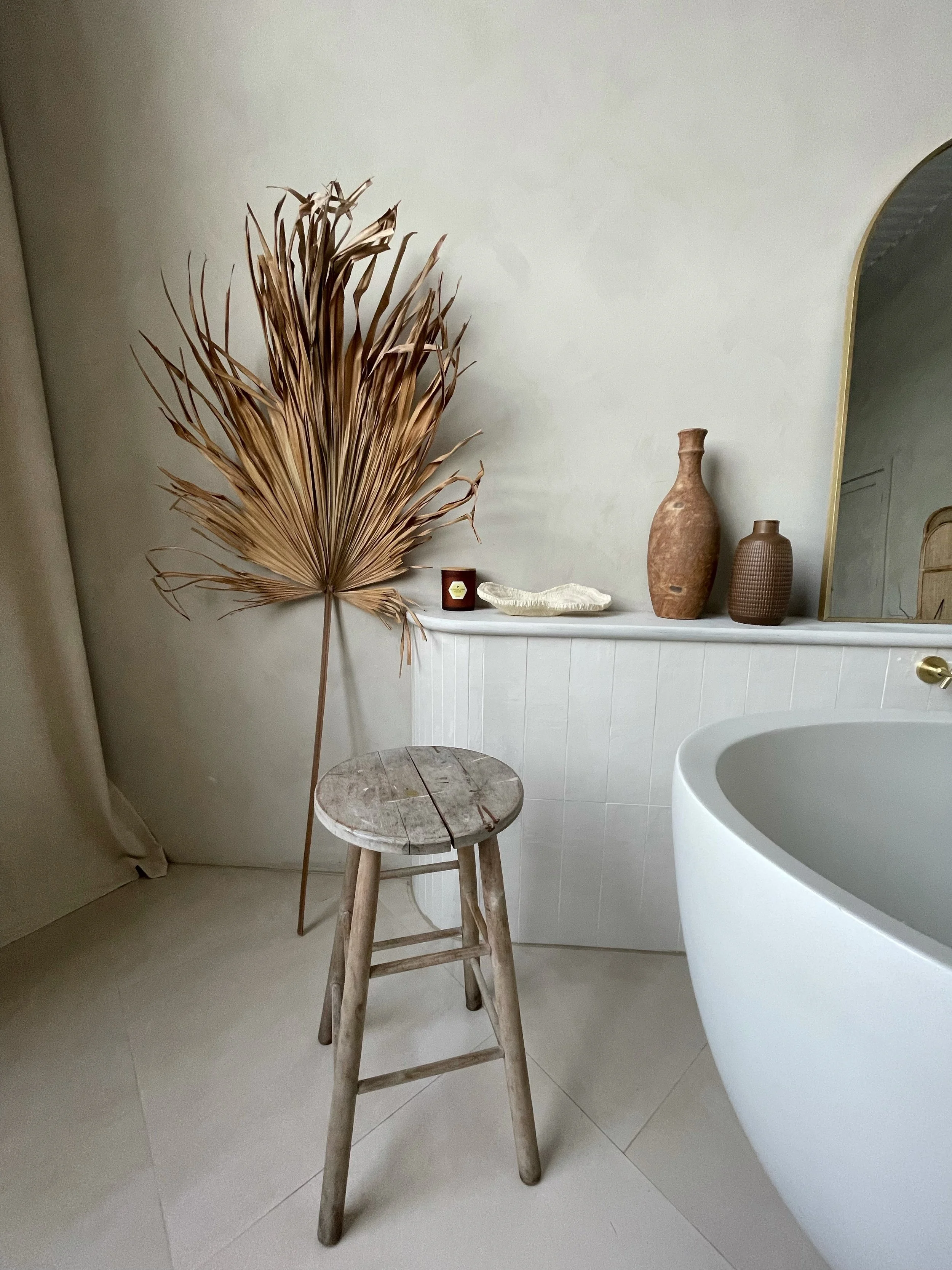 A bathroom corner with a wooden stool, large dried palm leaf, decorative vases, candle, and tray, with a curved bathtub and a mirror partially visible.
