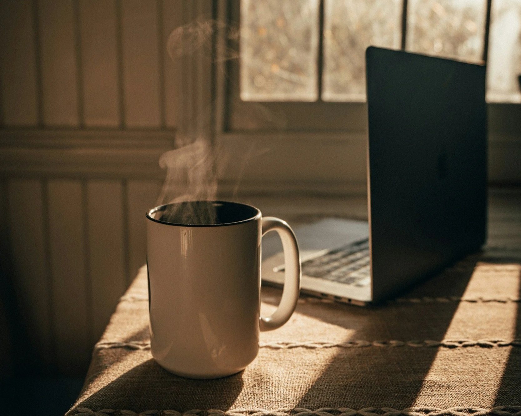 A steaming cup of coffee on a sunlit wooden table with a laptop in the background.