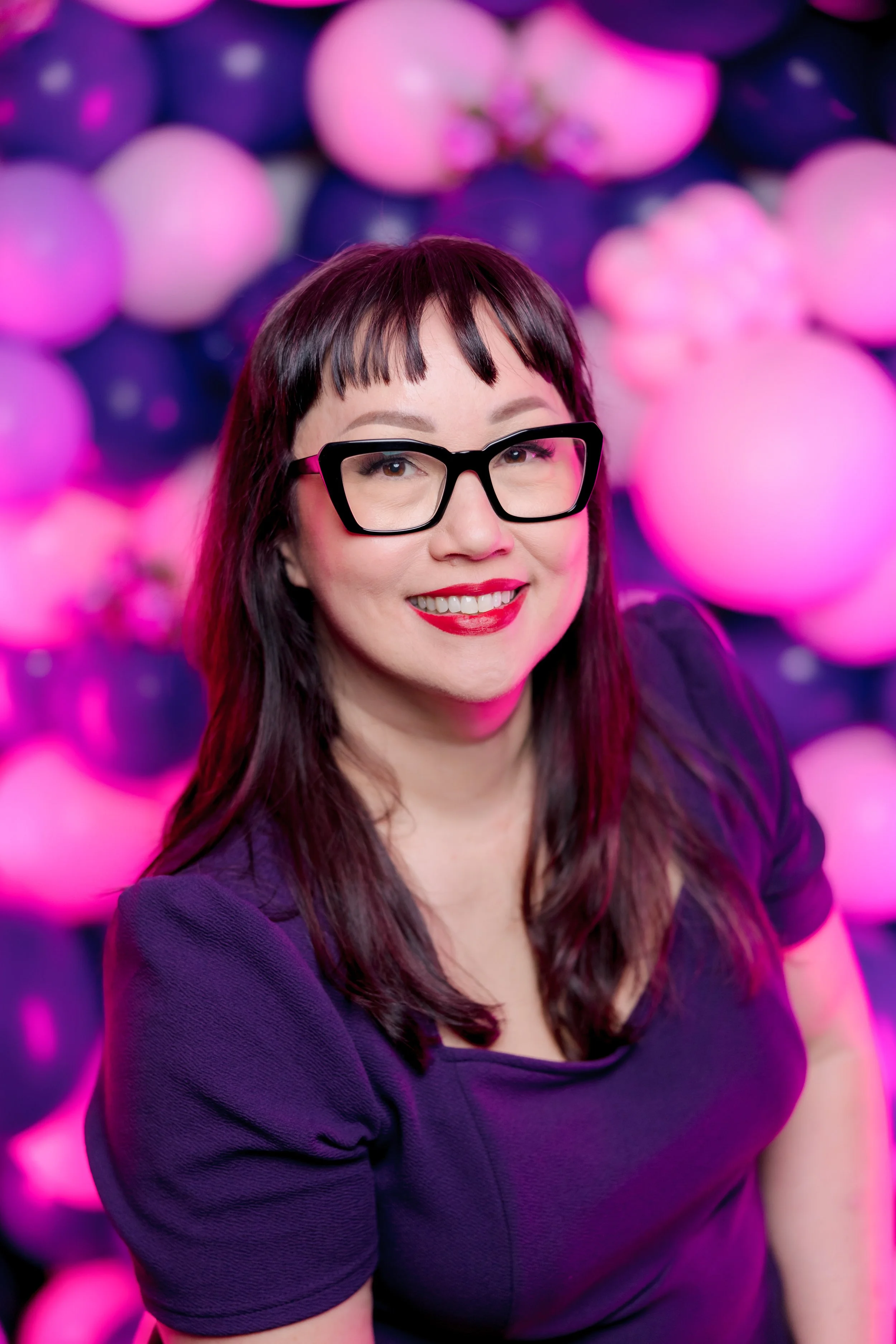 A woman with long dark hair, glasses, red lipstick, and a purple top, smiling in front of a background of pink and dark purple balloons.