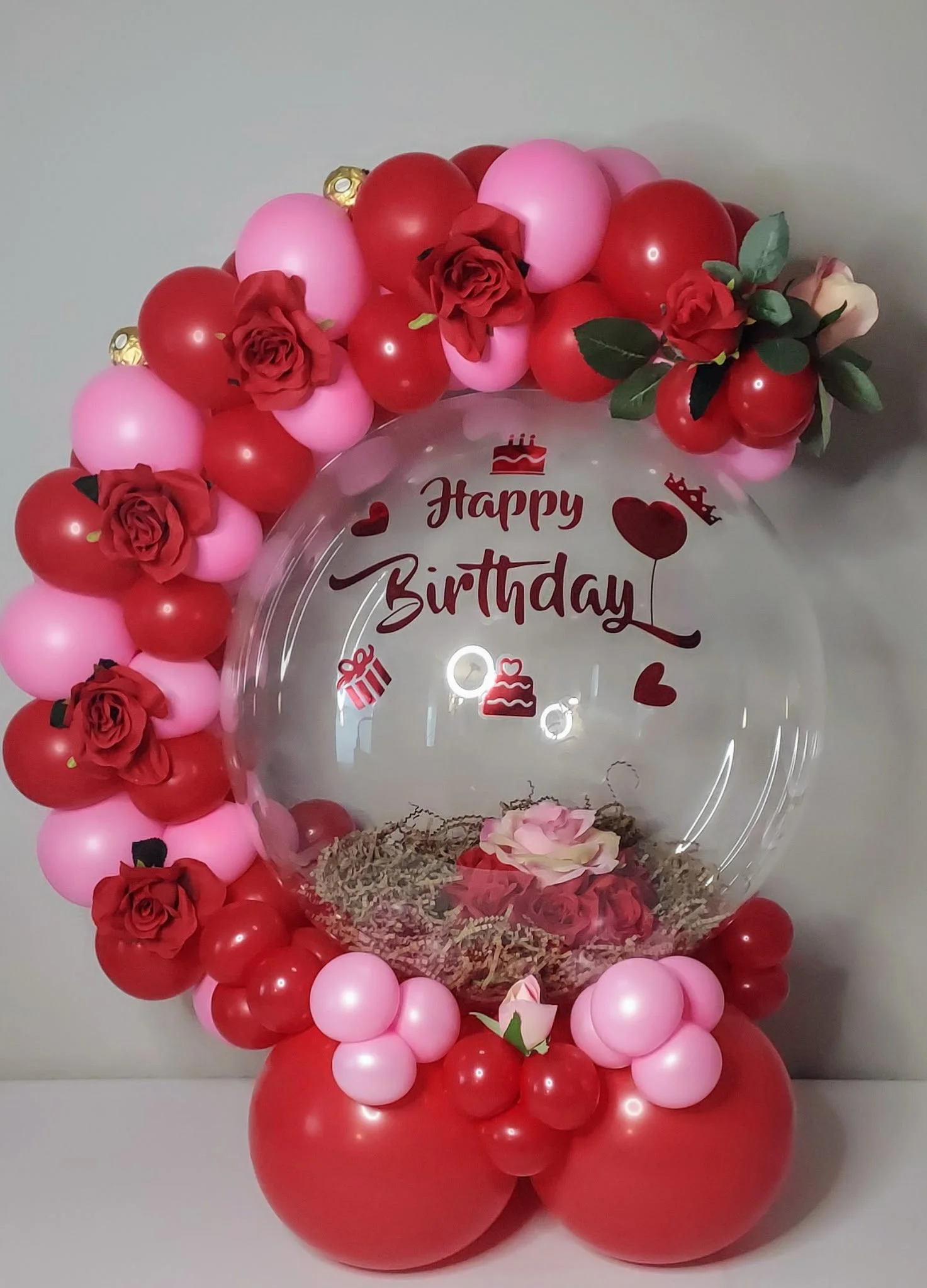 Decorative birthday display with pink and red balloons, faux roses, and a clear ball in the center with 'Happy Birthday' written on it, containing additional small decorative elements and a rose inside.