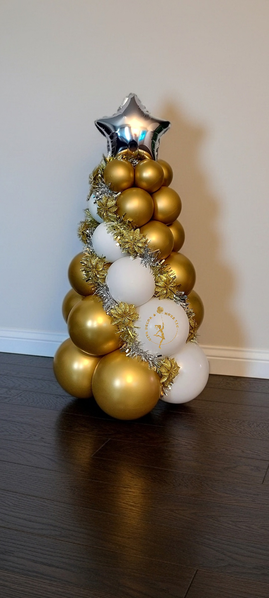 Christmas tree made of gold and white balloons, topped with a silver star balloon, decorated with gold and silver tinsel and gold poinsettia ornaments, with a logo on one of the white balloons.