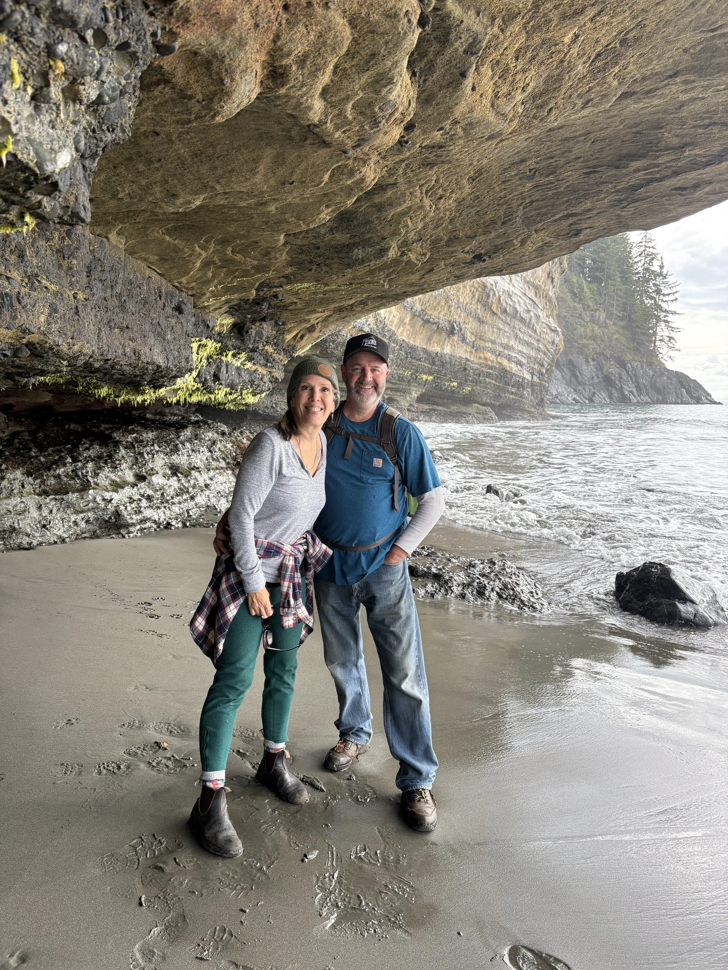 Two people standing on a sandy beach under a large rock formation, smiling and posing for a photo with the ocean and rocky cliffs in the background.