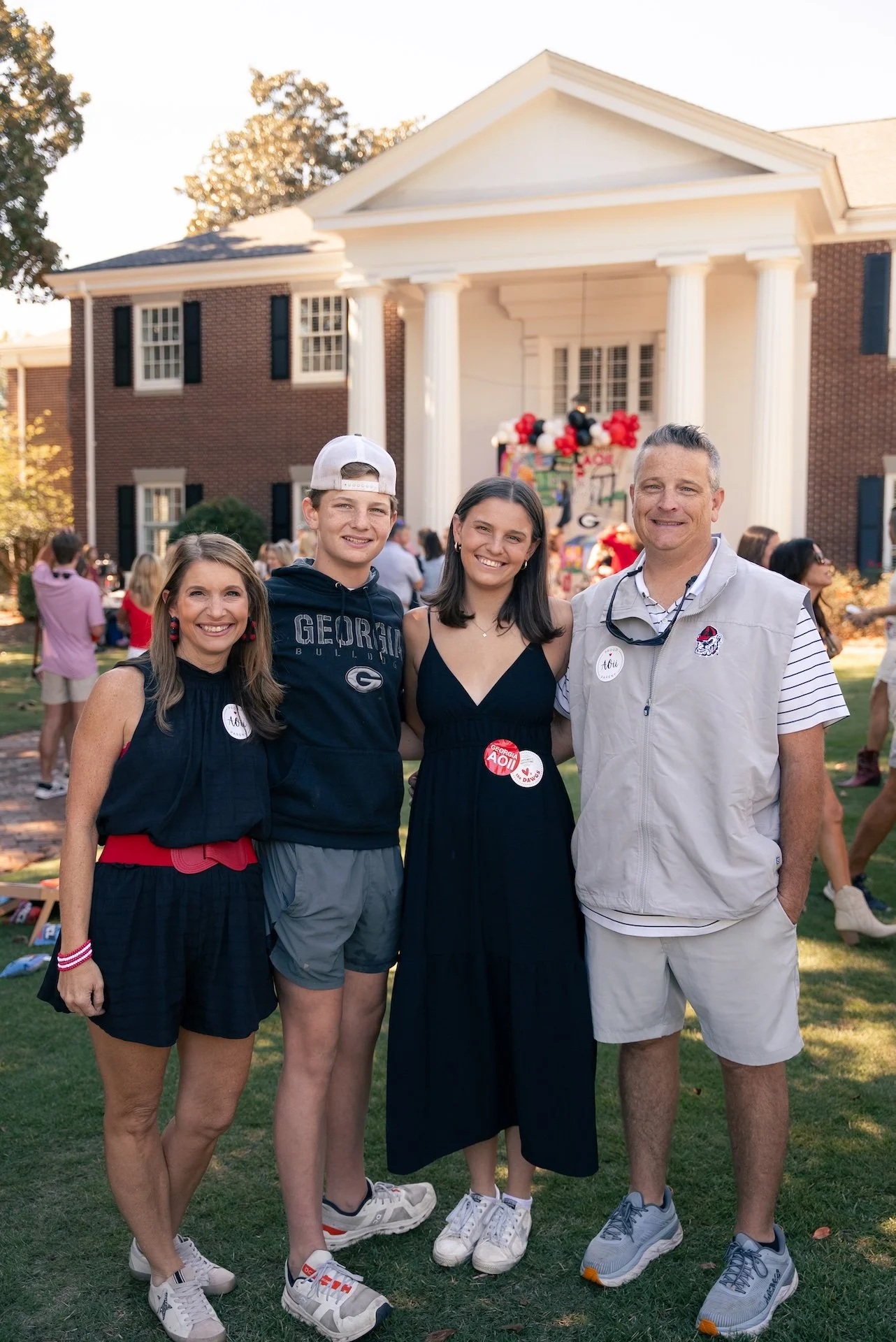 Family gathering outside a brick house with white columns, people in the background celebrating, some wearing Georgia Bulldogs attire, smiling and posing for a group photo.