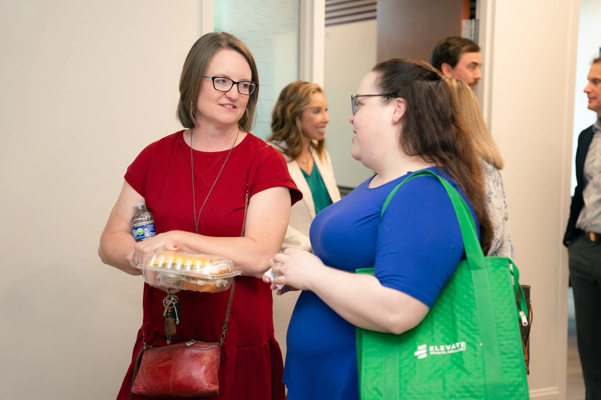 Two women having a conversation at an indoor event, with other people in the background.