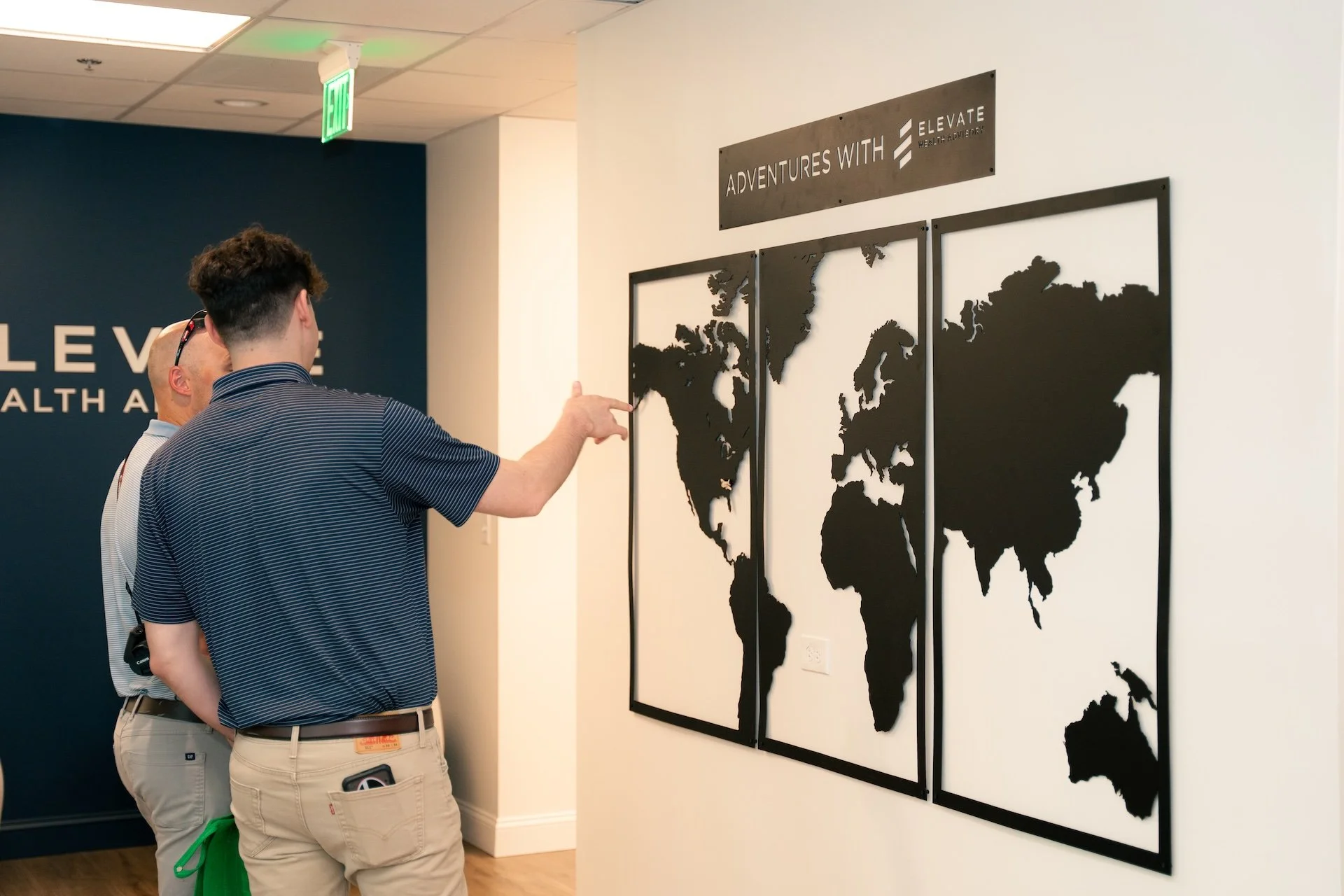 Two men looking at a black and white world map display in an indoor office setting. One man is pointing at North America.