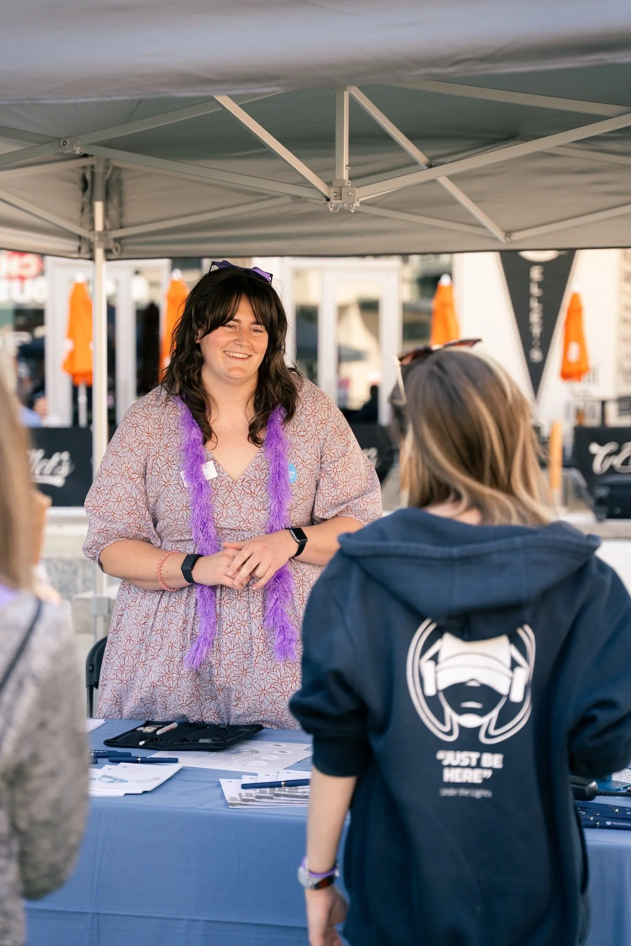 A woman with dark hair and a purple feathered accessory talking to a young girl at an outdoor booth under a tent, with other people and tents visible in the background.
