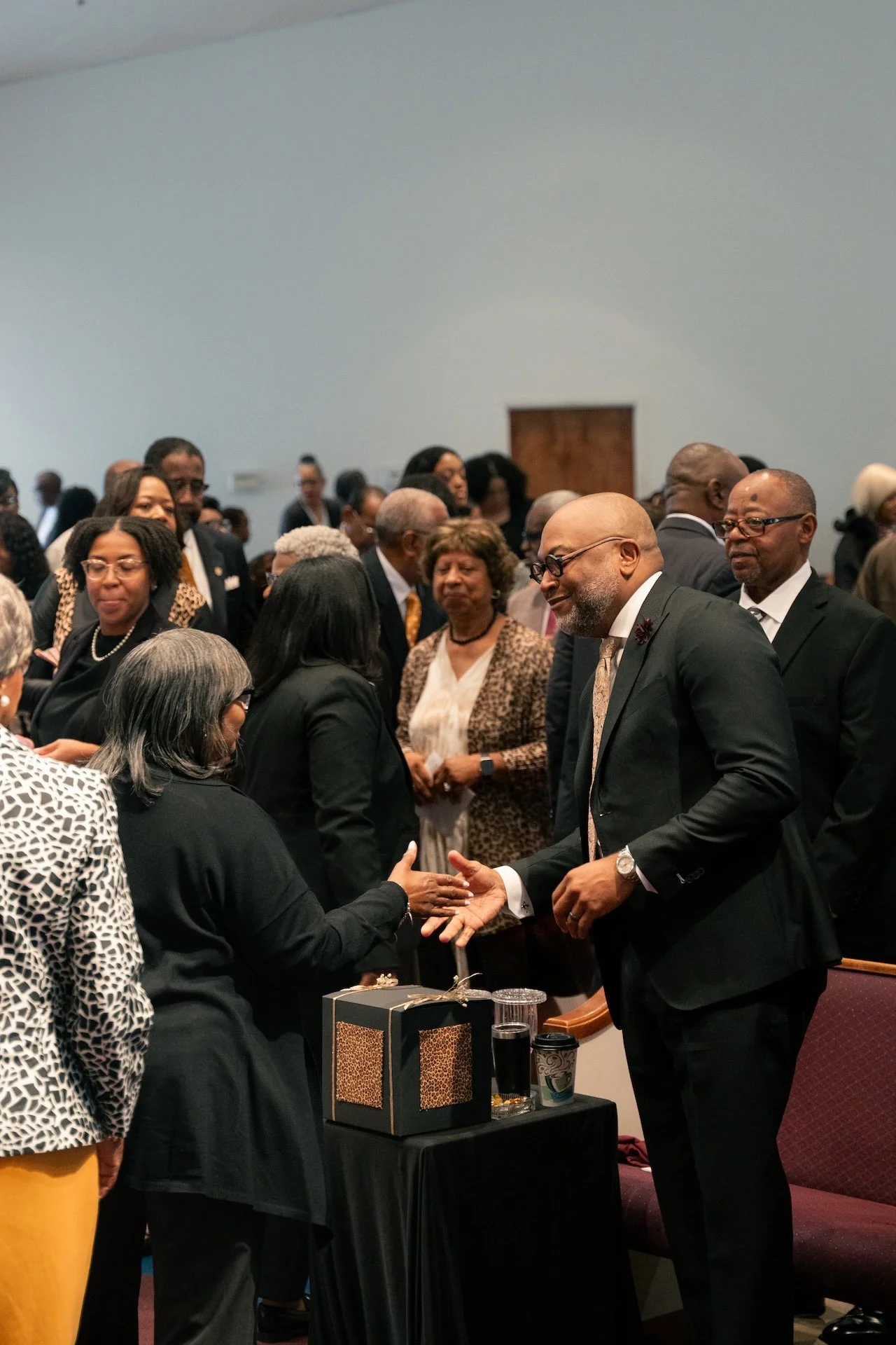 A man in a black suit shaking hands with a woman during a formal gathering or ceremony, with other people mingling in the background.