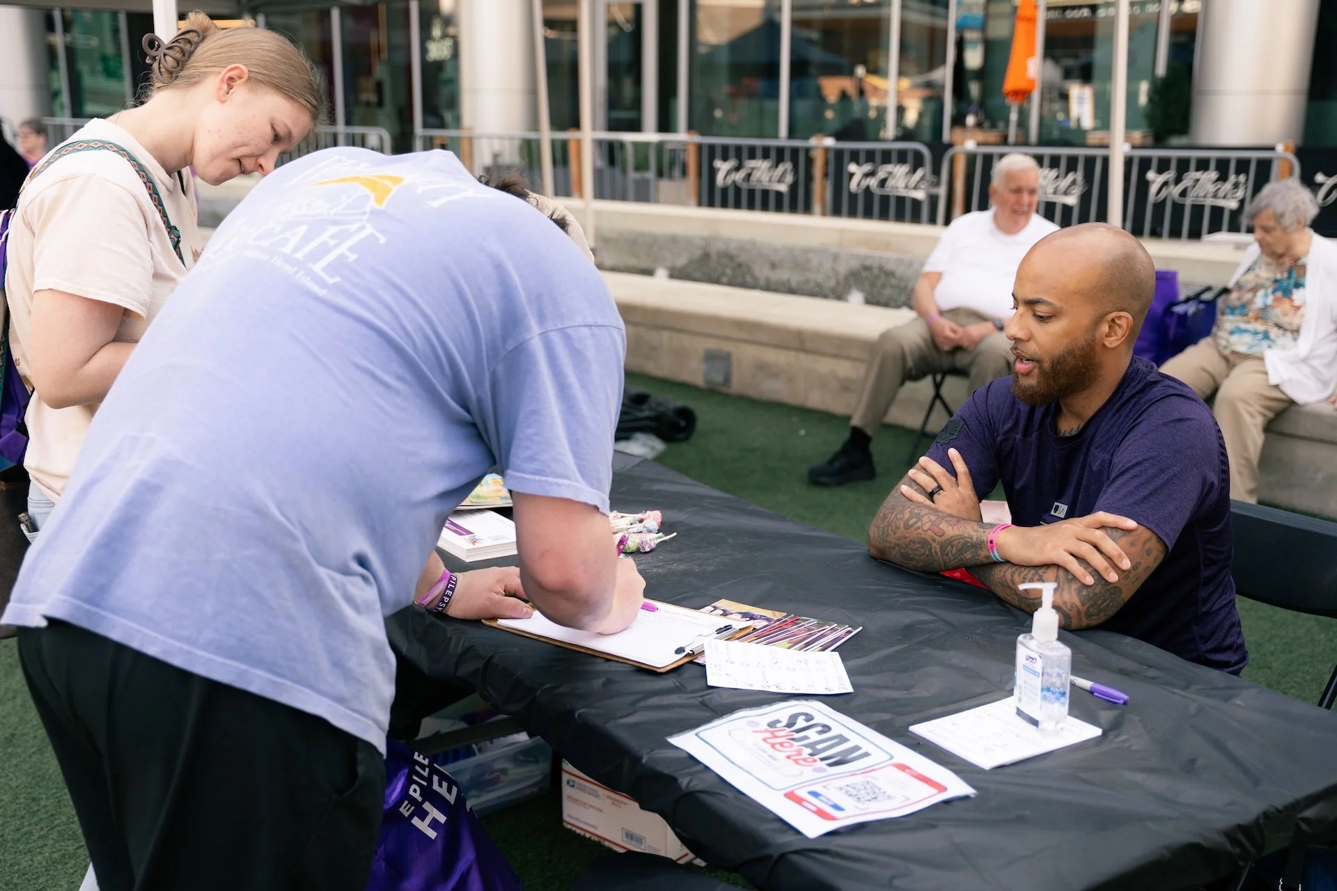 A young man with a shaved head and tattoos on his arms sitting at a table, talking to a woman and man standing in front of him; two women seated in the background watching.