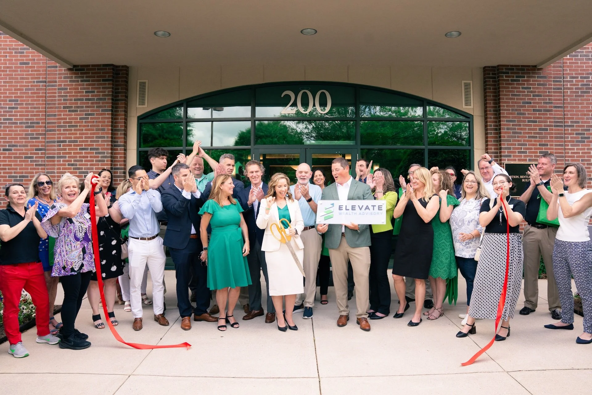 Group of people celebrating outside a building with a large group holding a sign that says Elevate Wealth Advisory, some holding scissors and a red ribbon, in front of the entrance marked 200.