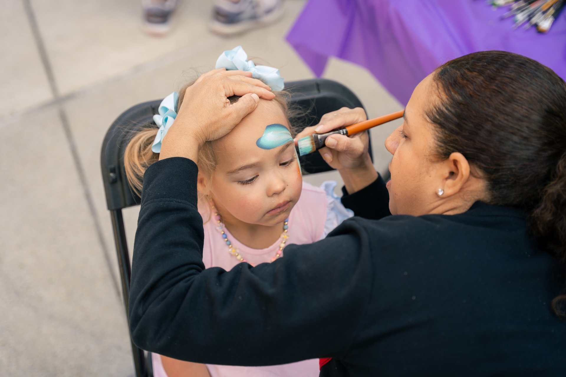 A woman is painting a blue cloud on a young girl's forehead with a brush at an outdoor event.