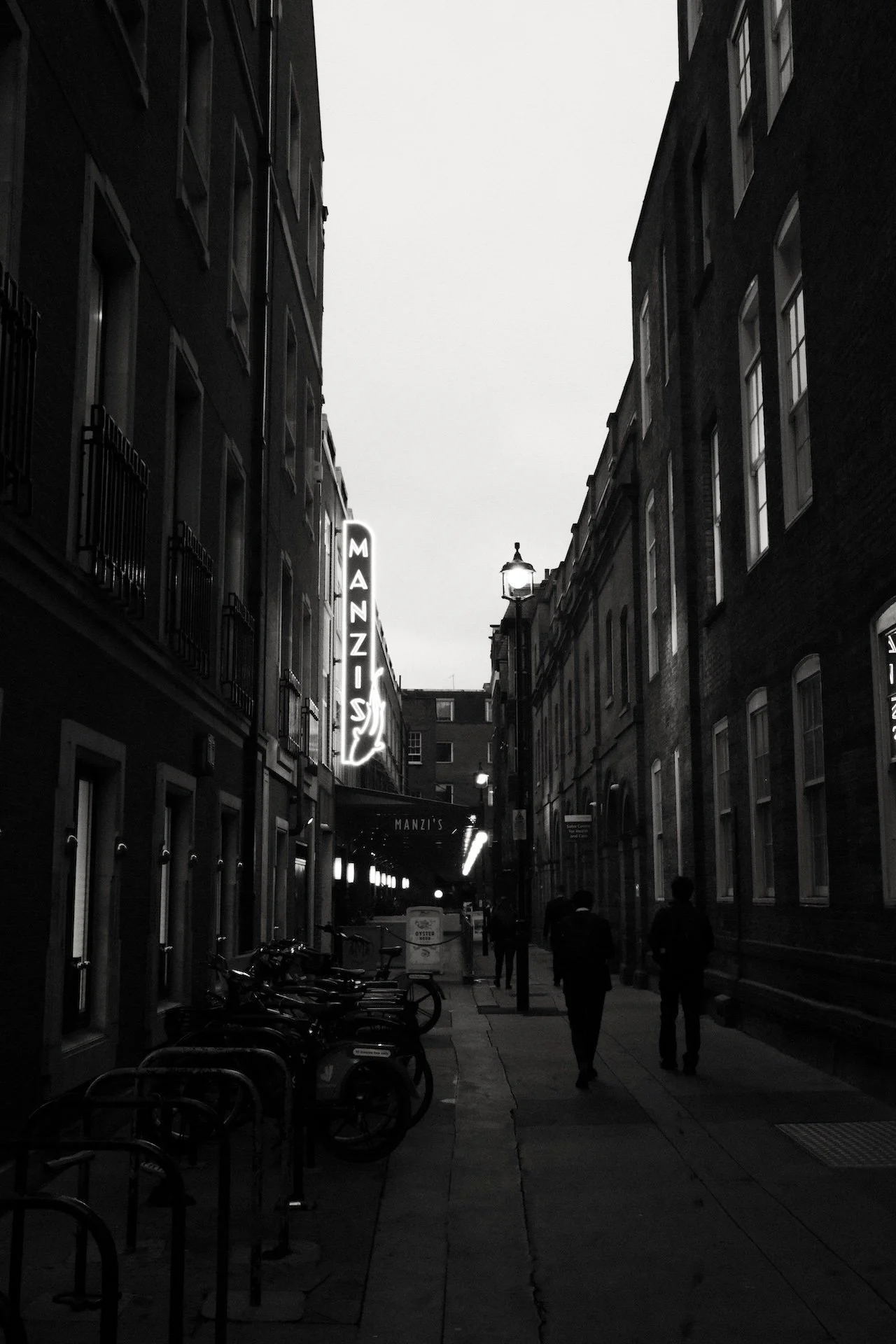 A narrow city street scene at dusk with a neon sign that reads 'MANZIS' hanging on the left side, bicycles parked along the sidewalk, and a few pedestrians walking under street lamps on both sides.