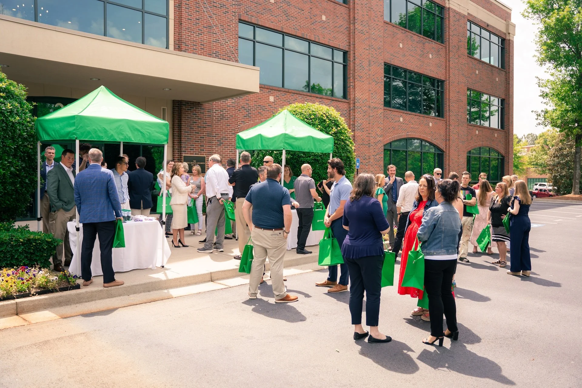 People gathered outside a brick building at a daytime event, under green tents, holding green bags, engaged in conversation.