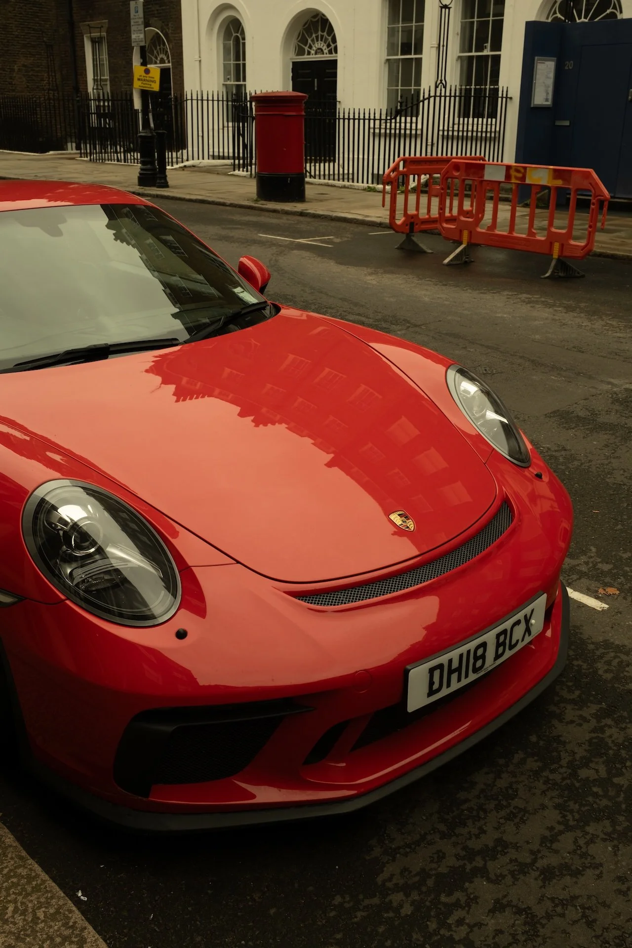 A red Porsche sports car parked on a city street with a white building and orange construction barriers in the background.