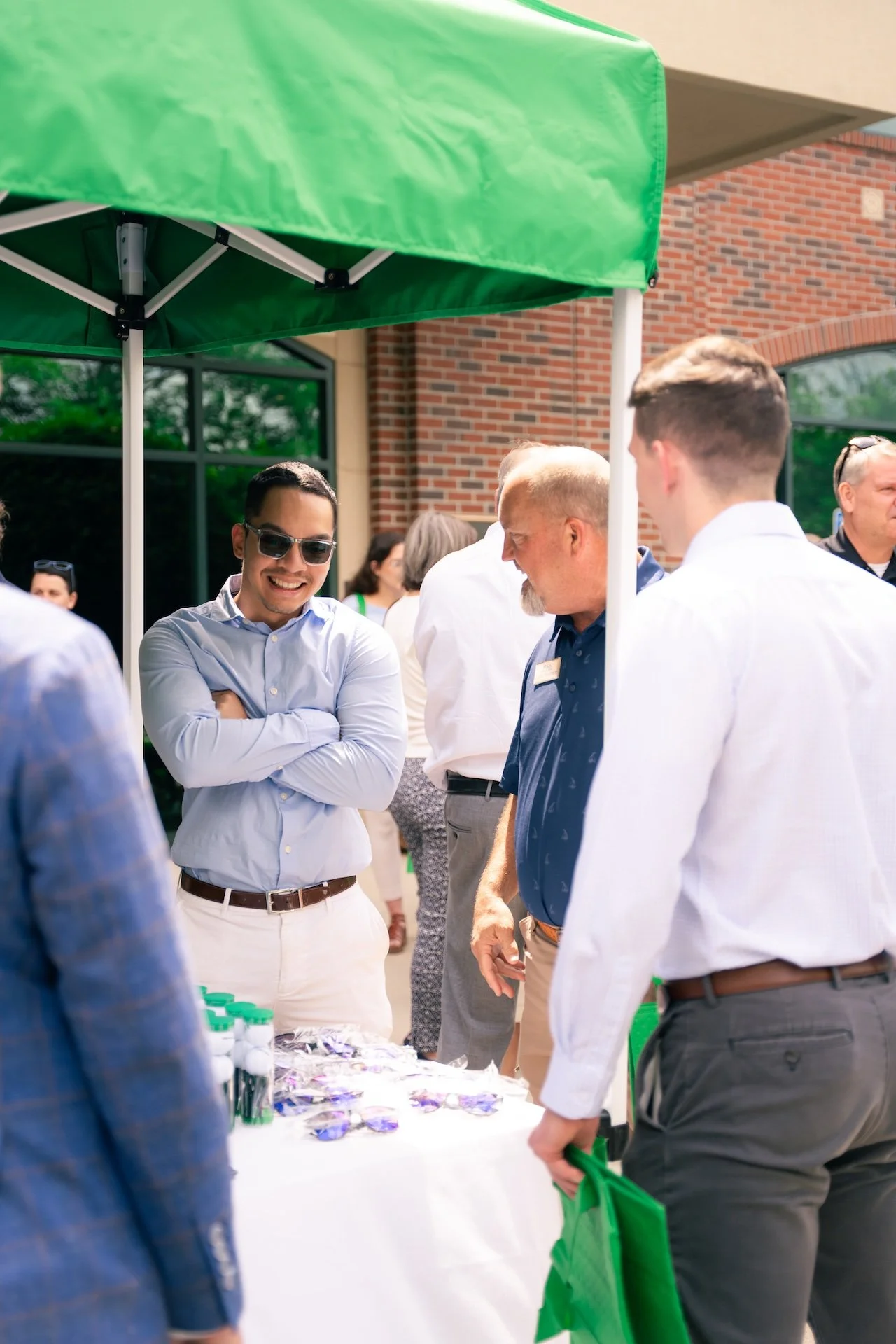 Group of people socializing outdoors at a table with promotional items under a green canopy.