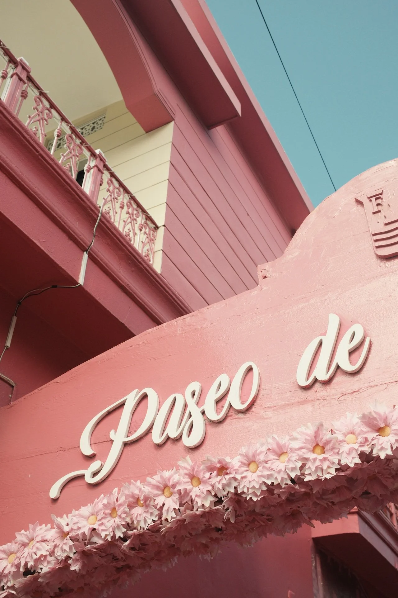 Pink storefront sign with white cursive text that reads 'paseo de', decorated with artificial pink flowers.