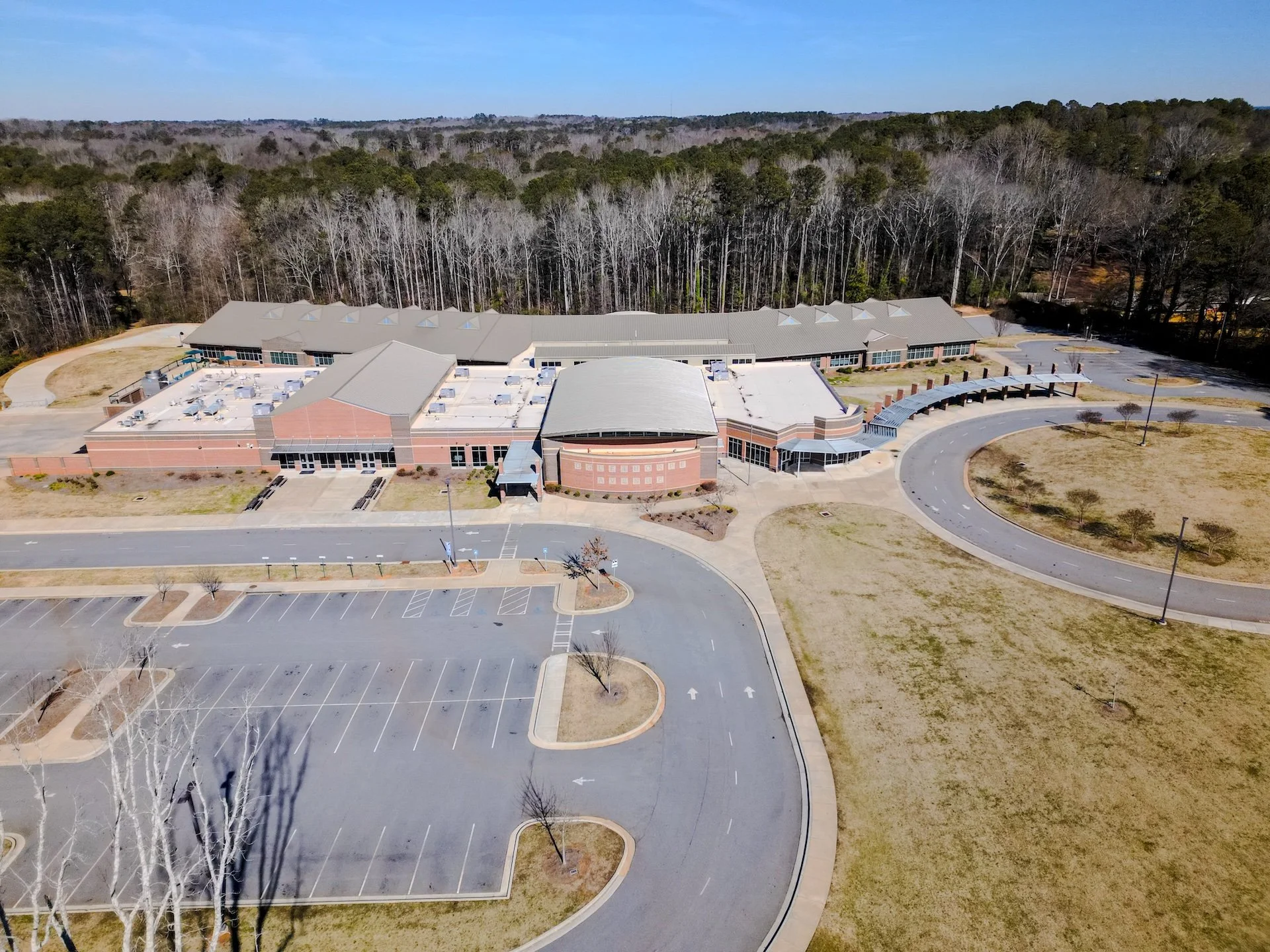An aerial view of a large, modern school building with a parking lot, surrounded by trees and grass on a clear day.