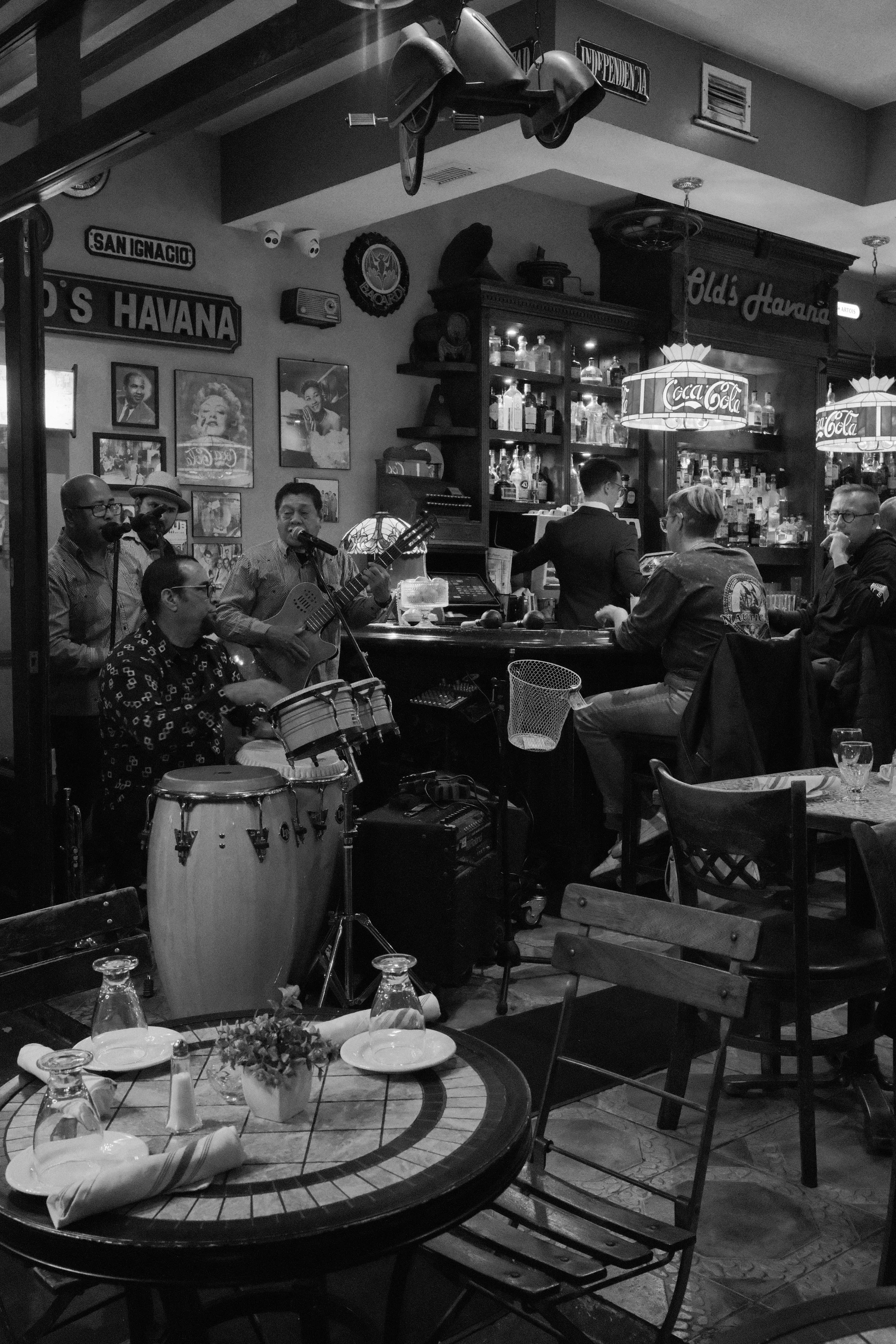 A band performing live music at a cozy restaurant or bar, with a drummer, guitarist, and singers, surrounded by patrons at tables, bar shelves with bottles, and vintage decor.