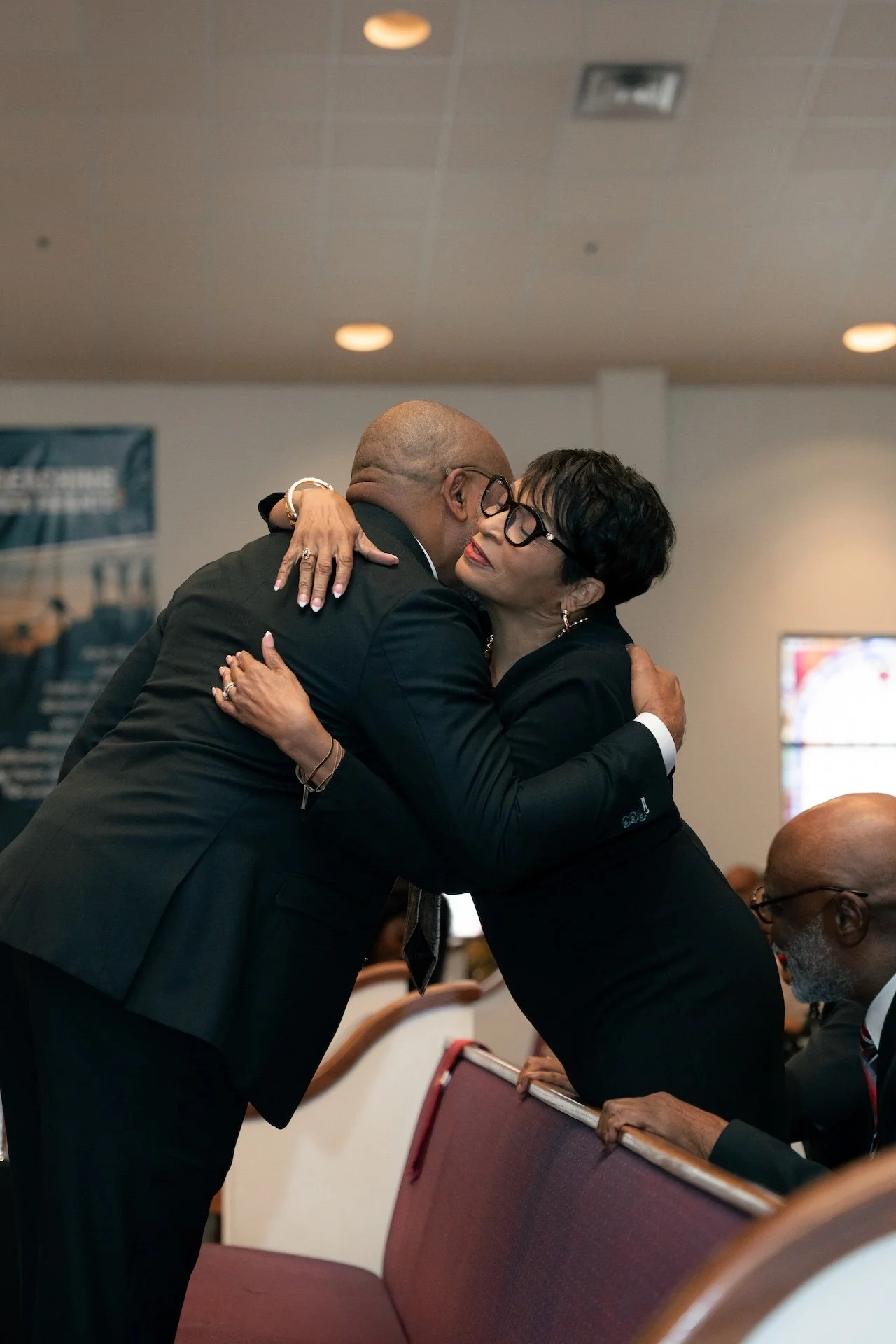 Two people embracing in a hug at a formal event, with one person standing and the other sitting, both dressed in dark clothing.