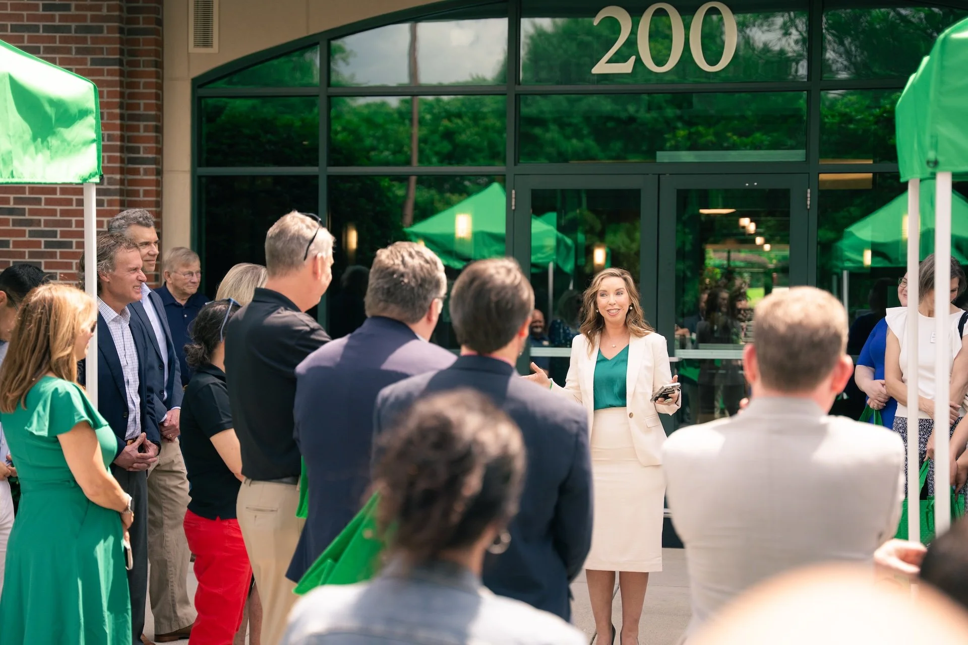 A woman in a white suit and green blouse speaking to a group of people outside a building with the number 200 on it. The crowd is standing under green umbrellas, and the group is listening attentively.