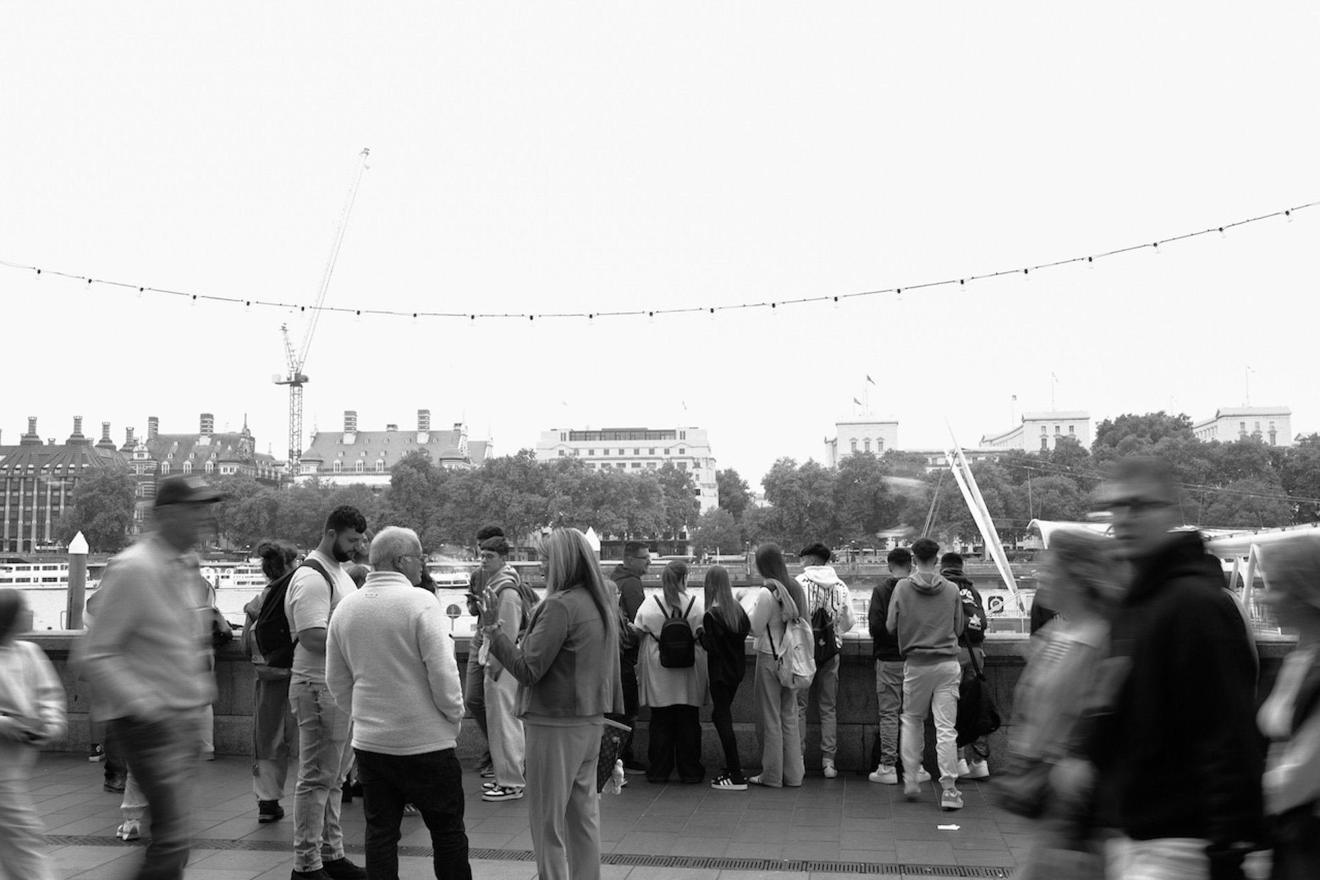 People walking and standing along a waterfront promenade with buildings and trees in the background, some blurred due to movement, in black and white.