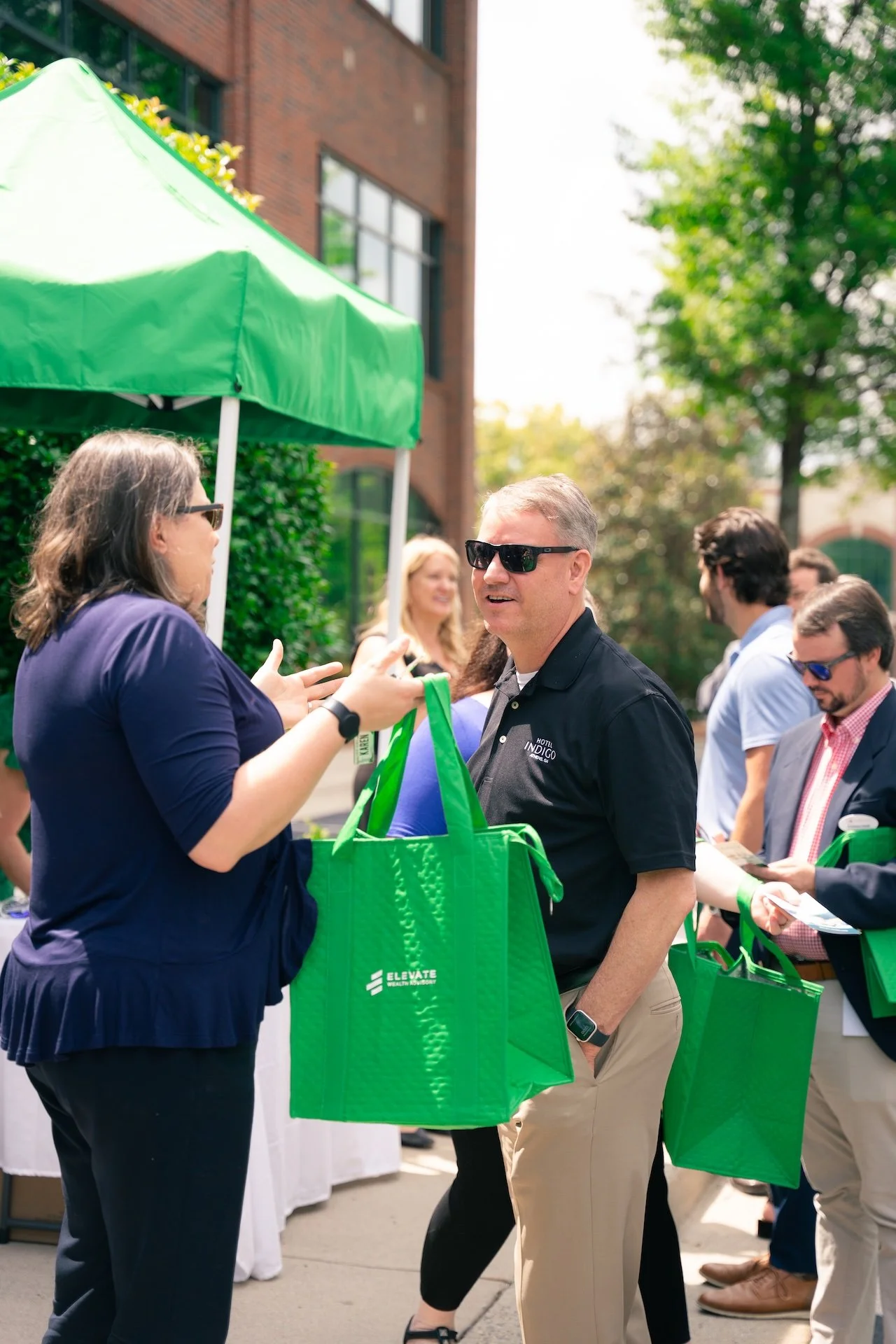 A woman and a man wearing sunglasses are having a conversation at an outdoor event with green tote bags. Several people are visible in the background, some holding green bags, under a green canopy with a brick building and trees nearby.