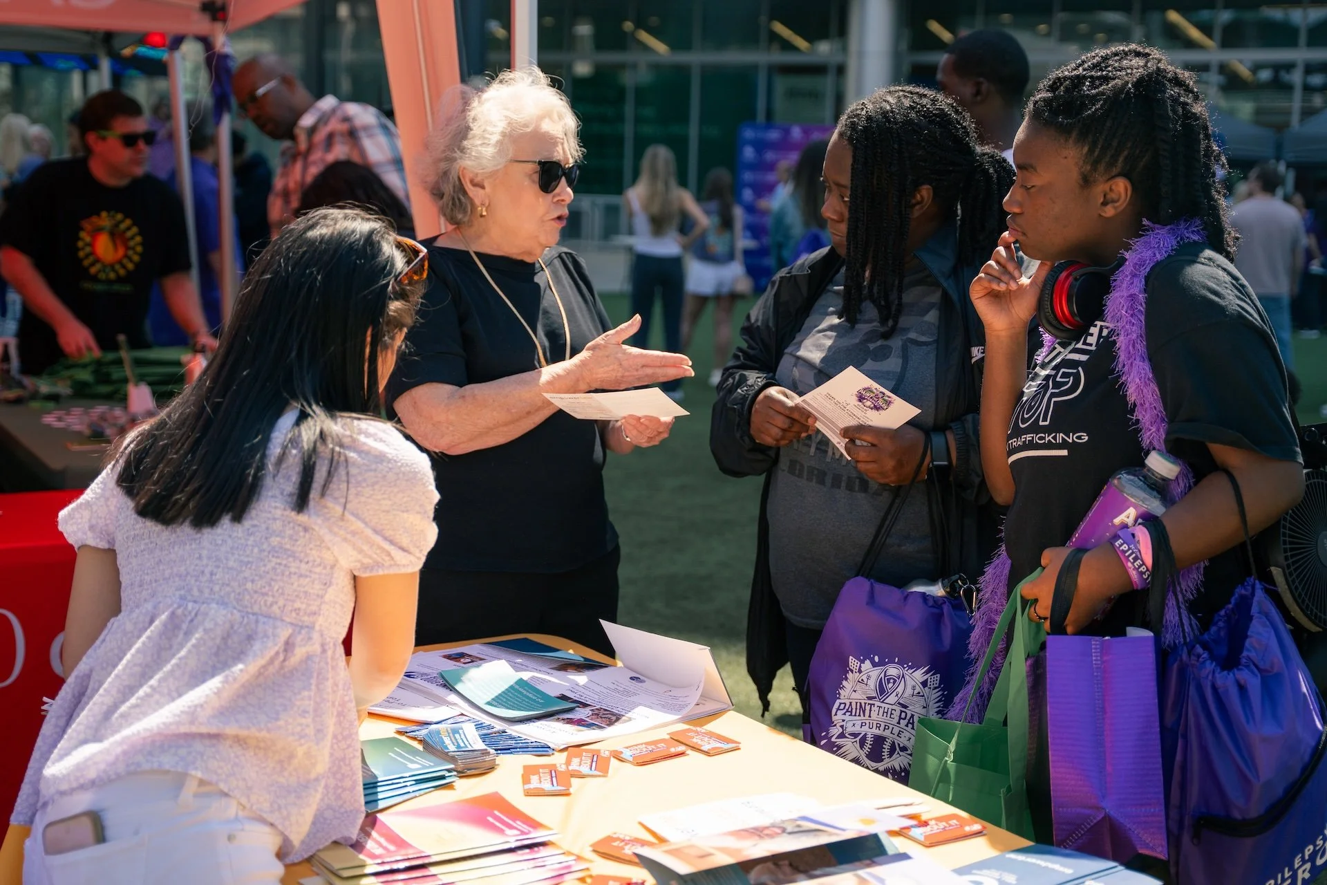 A group of people gathered around an informational booth at an outdoor event, listening to a woman with short white hair and sunglasses explain something to three women of different ethnicities. The booth has flyers and pamphlets on the table.