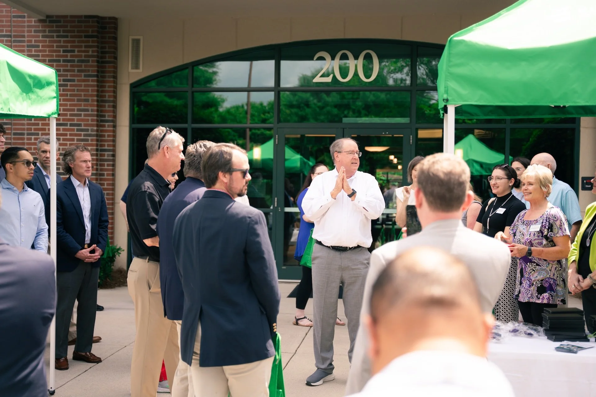 A group of people gathered outside a building with the number 200 on it, listening to a man speaking with his hands clasped in a prayer-like gesture. Green tents are set up around them.
